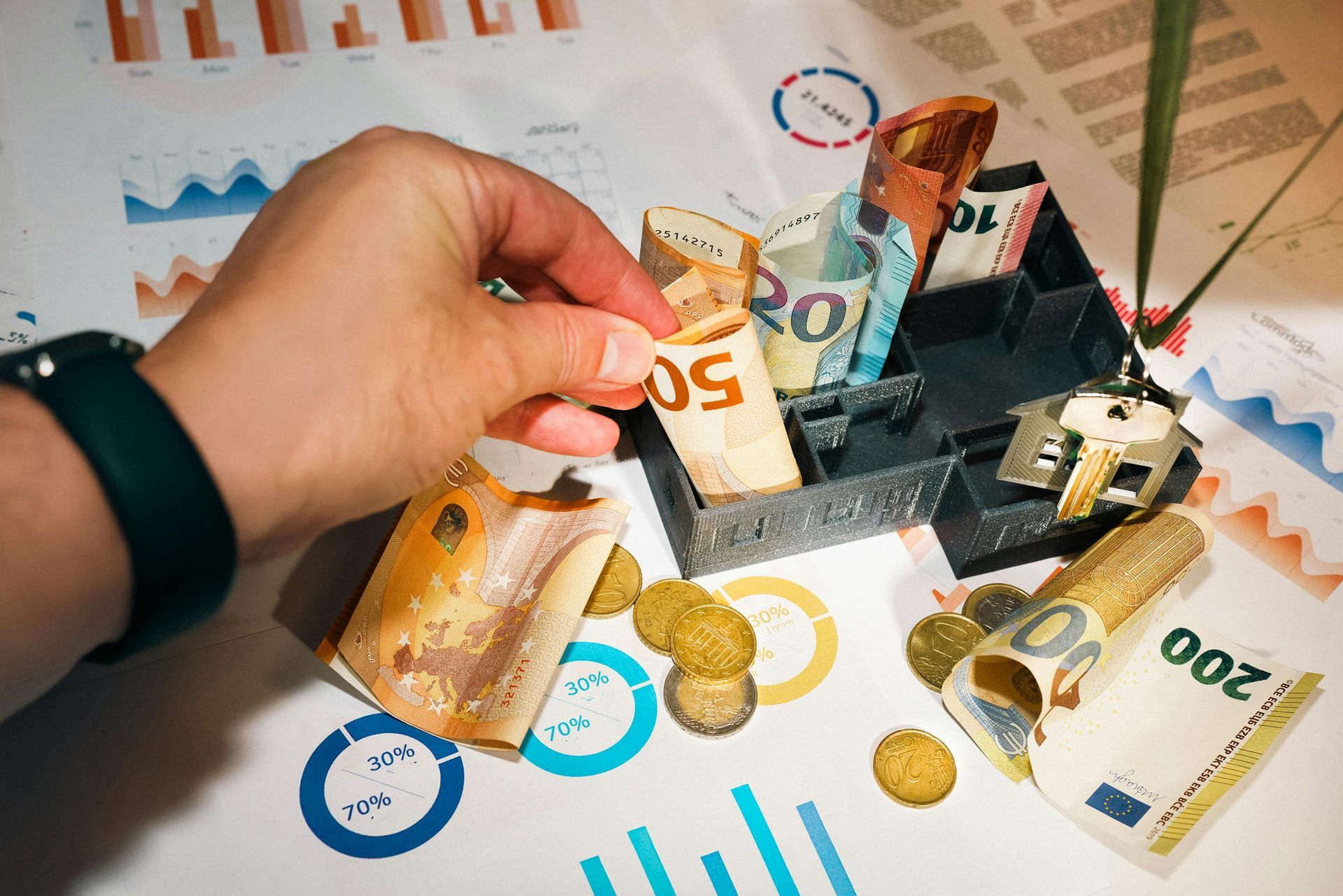 Person's hand placing a 50 Euro note in a black cash holder with other cash and coins scattered on charts.