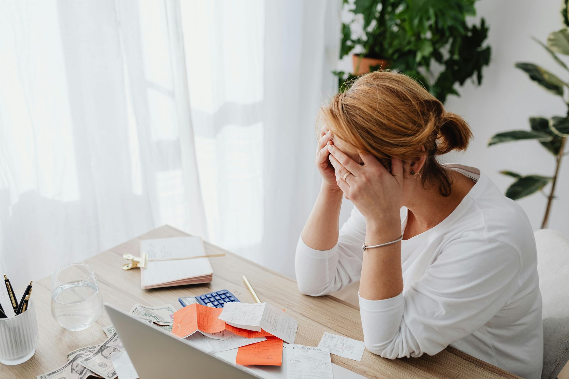 Woman with hands on her face, sitting at a desk with bills and laptop, appearing stressed.