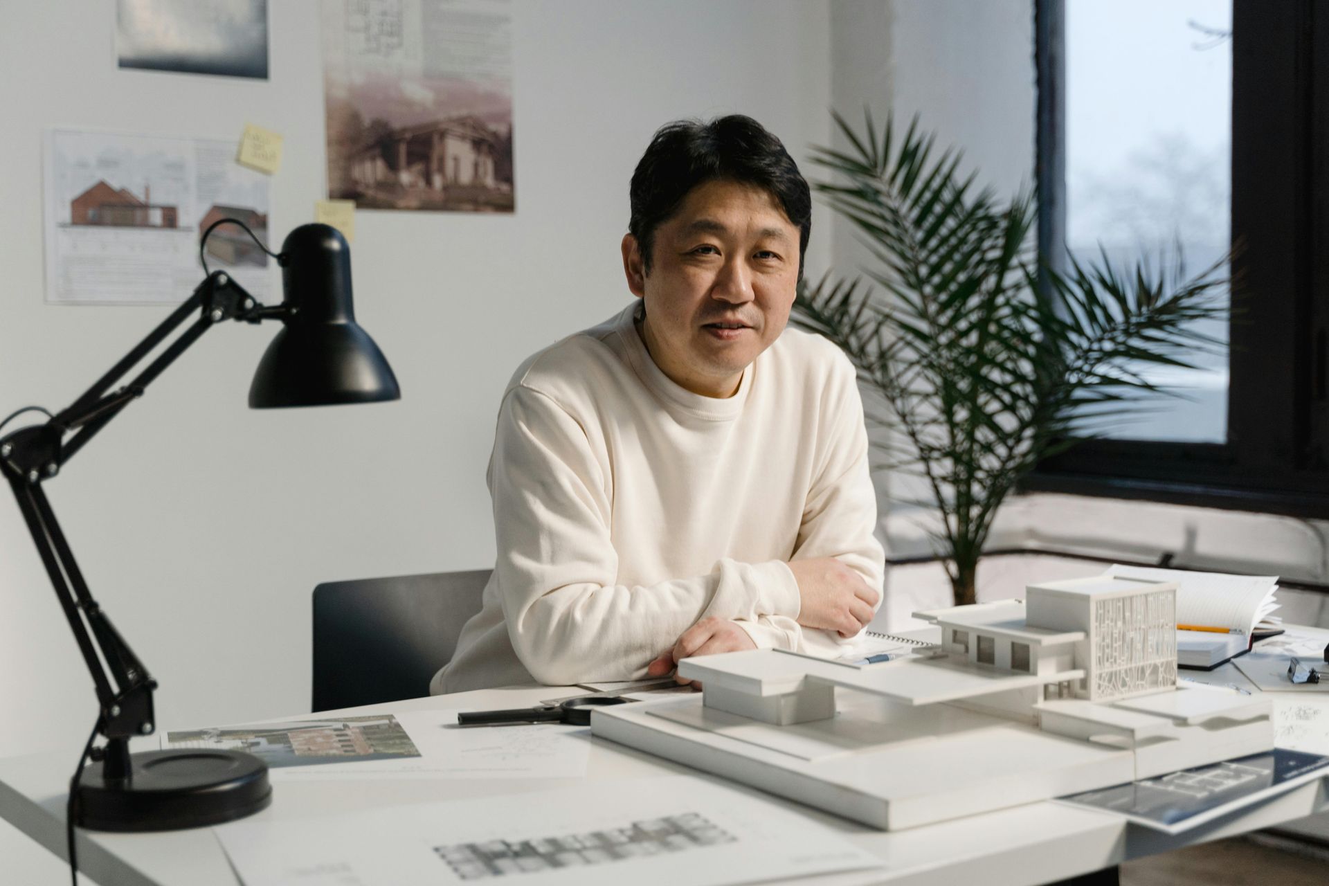 Man sitting at desk with architectural model, plant, and lamp. White sweater, smiling, appears to be an architect.