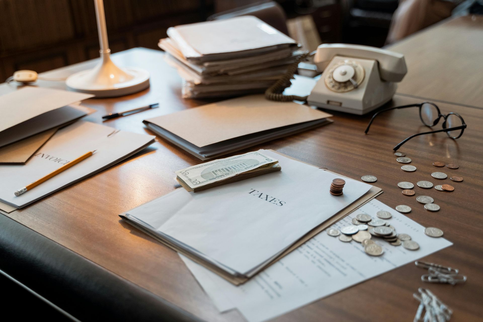 Desk with documents, stacked papers, vintage rotary phone, glasses, and scattered coins in warm light