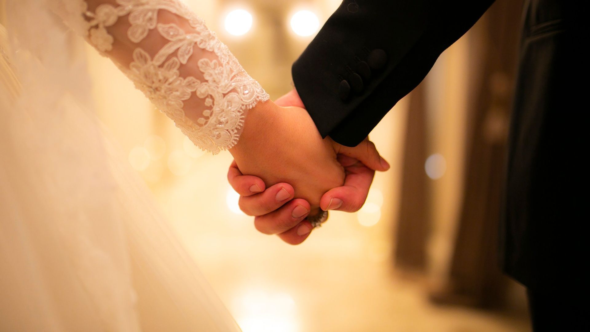 Bride and groom holding hands, focus on clasped hands with lace sleeve and suit.