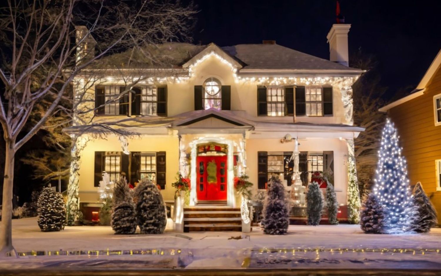 Festive house decorated with white lights, red door, and snow-covered yard at night.
