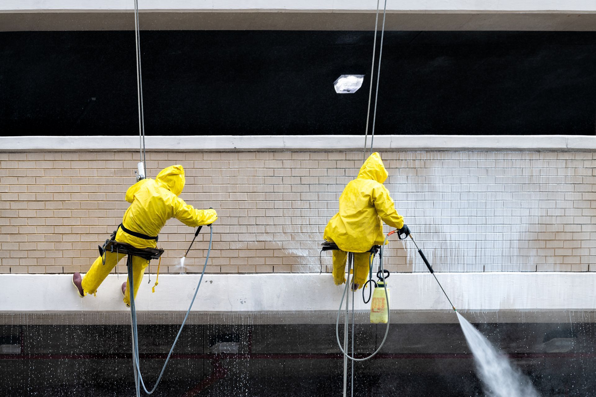 Two men in yellow jackets are cleaning a brick wall with a high pressure washer.
