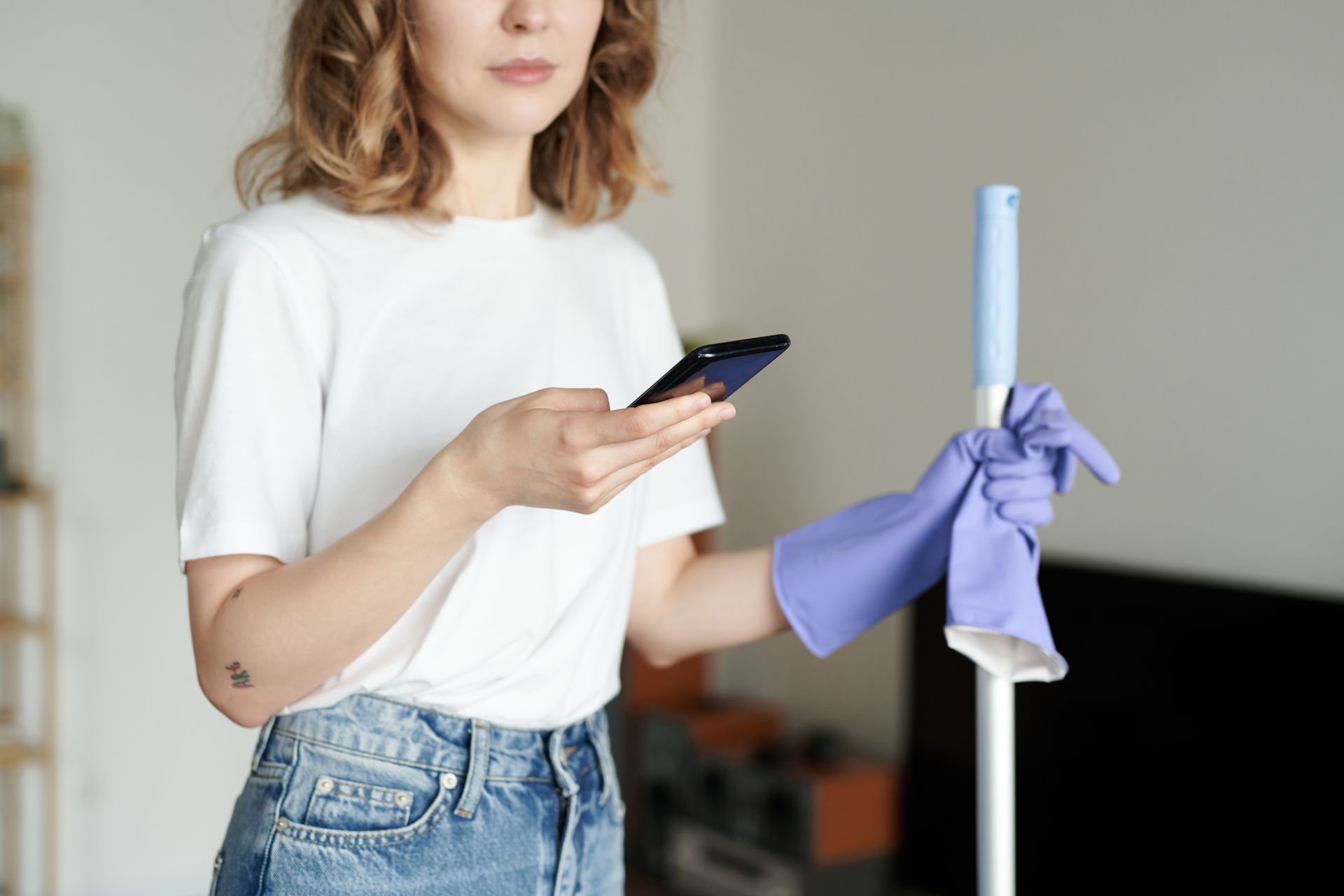 A woman is holding a mop and a cell phone.