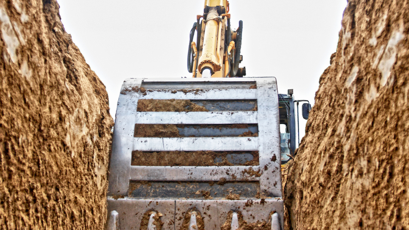 Excavator bucket in a trench, viewed from inside the ditch, with dirt walls and a light sky.