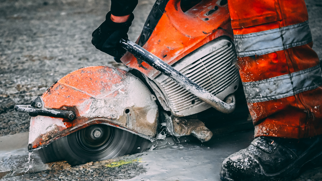 Person in orange workwear uses a concrete saw, creating dust.