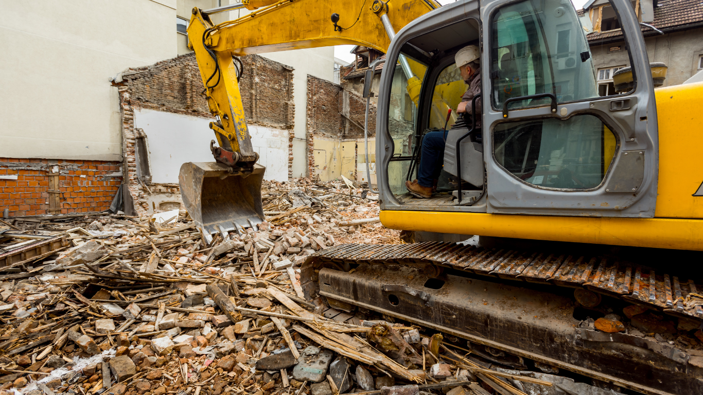 Yellow excavator demolishing a building; operator inside. Debris and exposed structure visible.