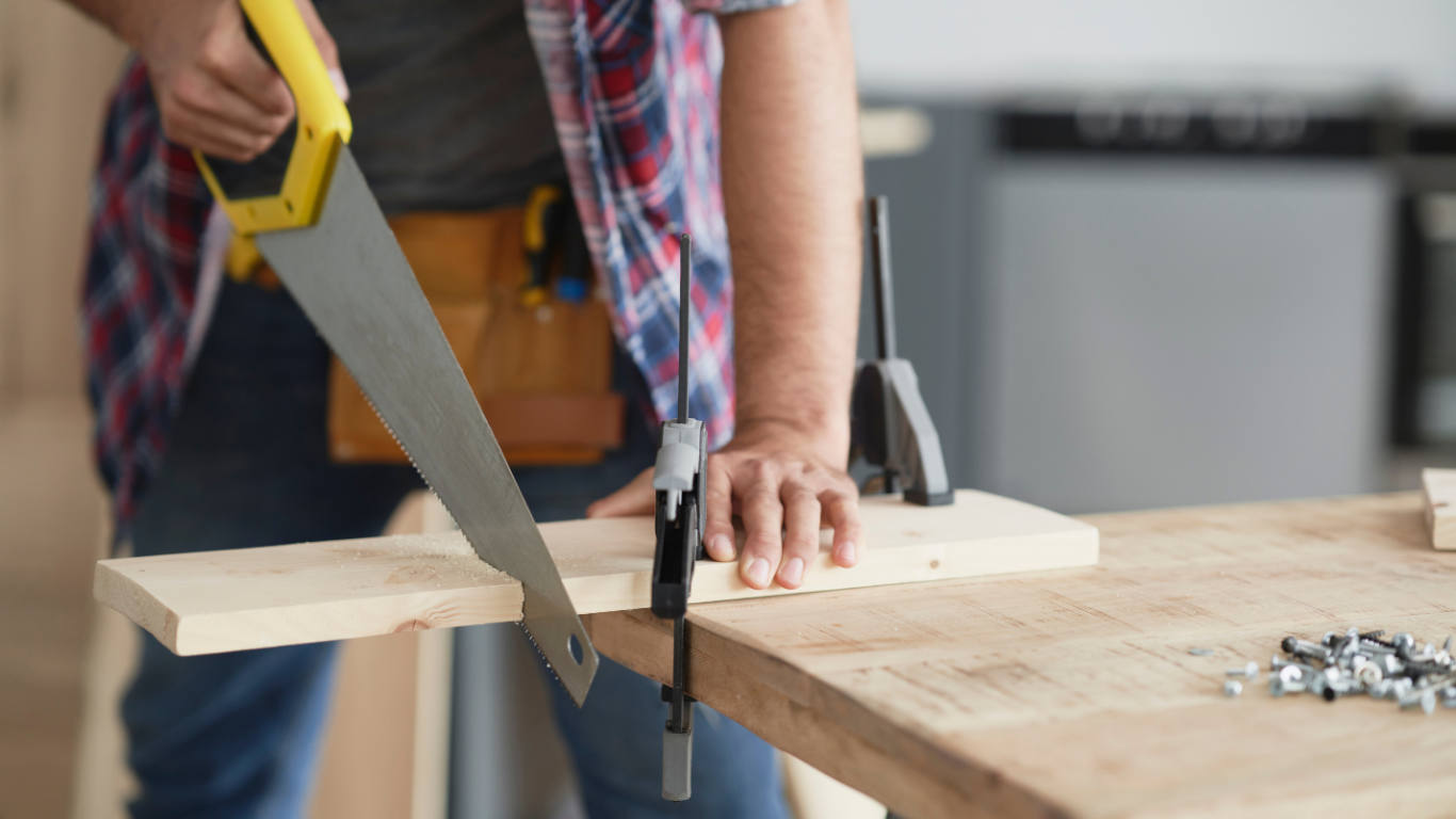 Person sawing a wooden board clamped to a workbench, wearing a plaid shirt.