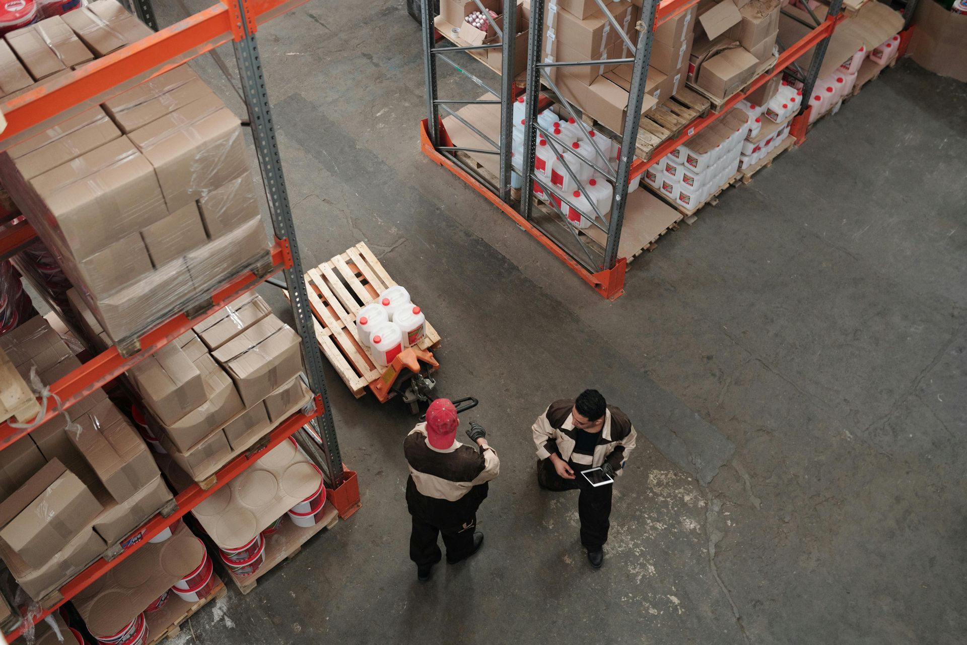 Warehouse scene: Two workers with a pallet jack, loading or unloading goods next to shelving filled with boxes.