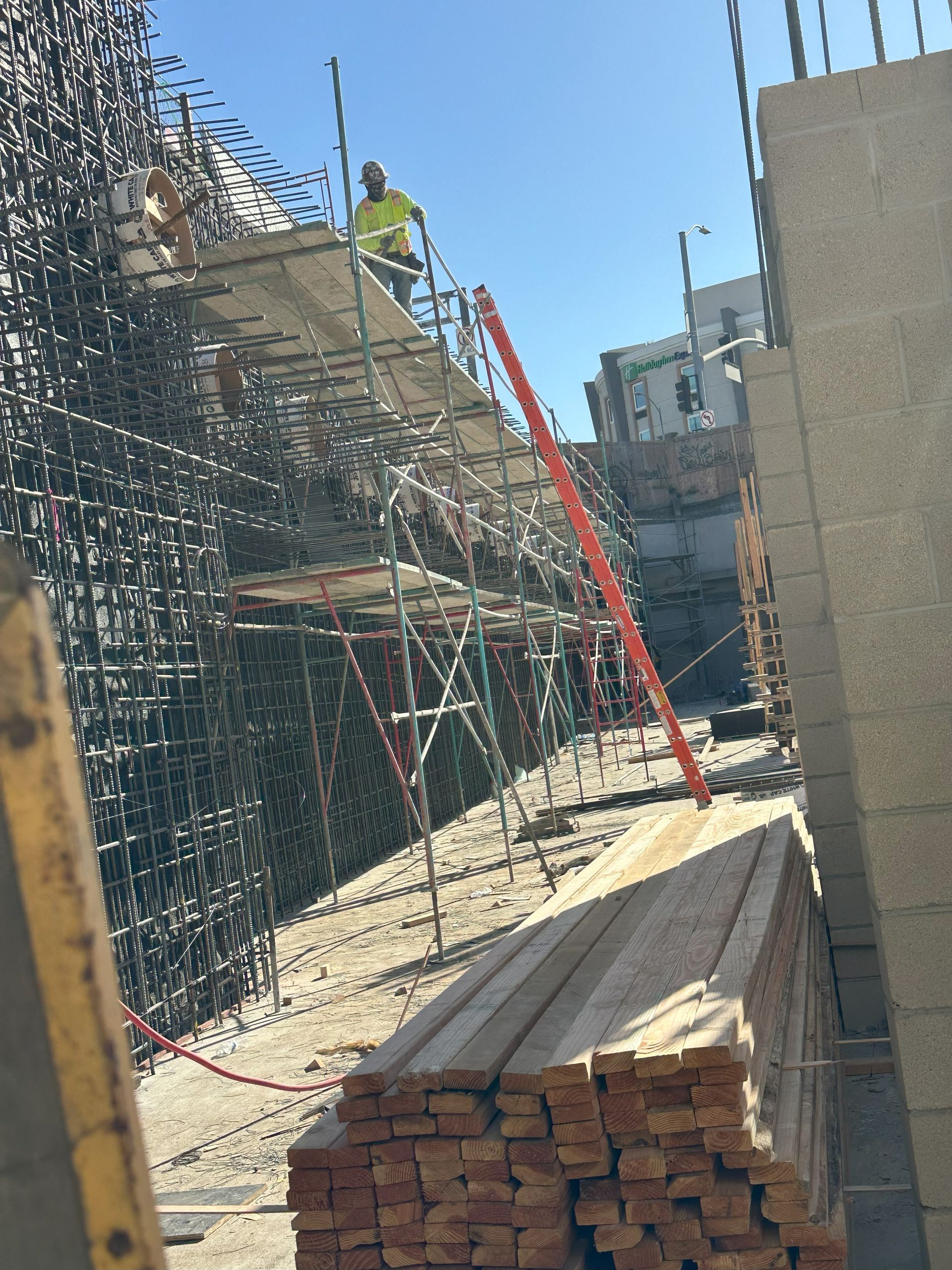 Construction worker on scaffolding, working next to a concrete wall, sunny day. Stack of wood in the foreground.