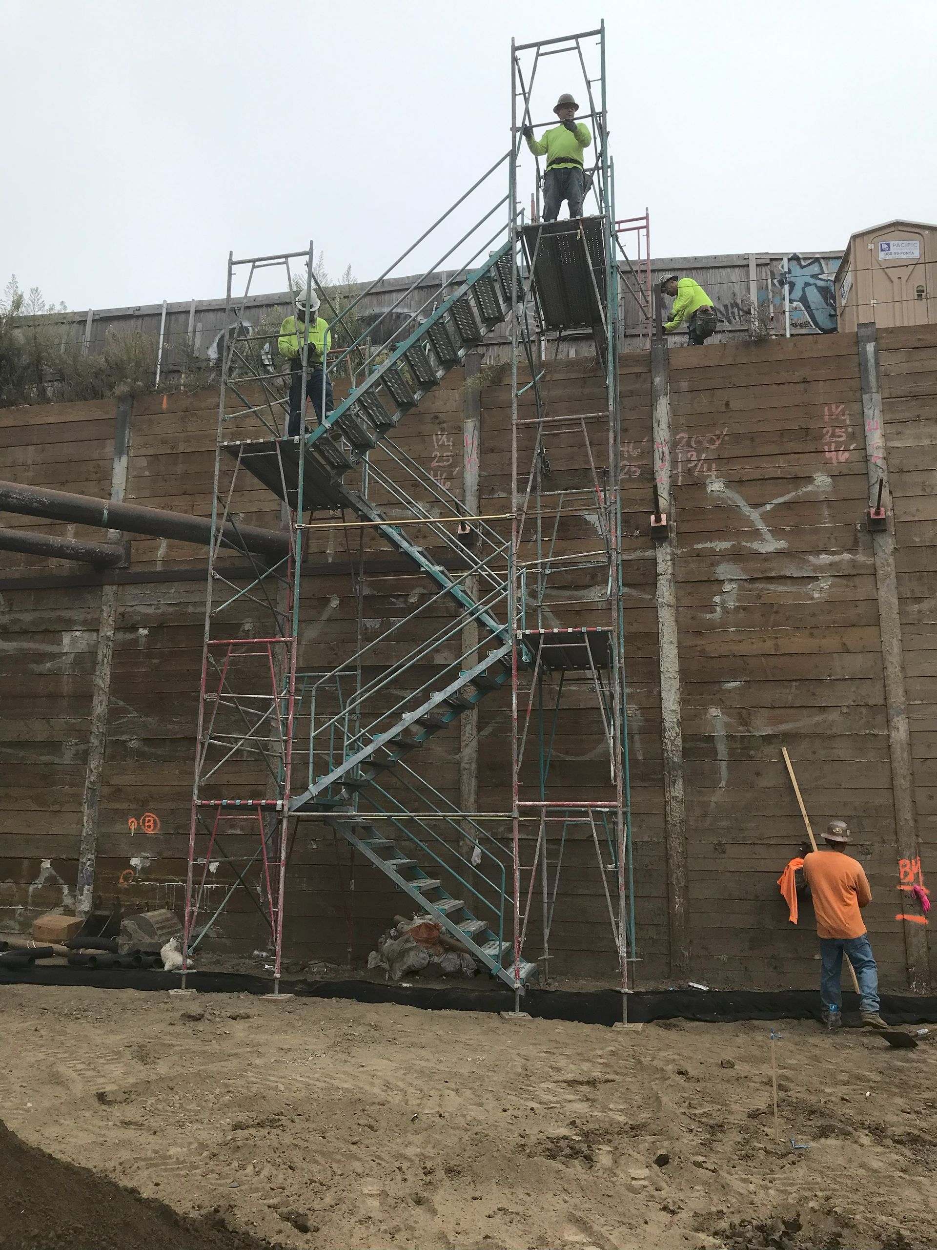 Construction workers on scaffolding against a weathered wooden wall. One worker shovels, others on the platforms. Cloudy day.