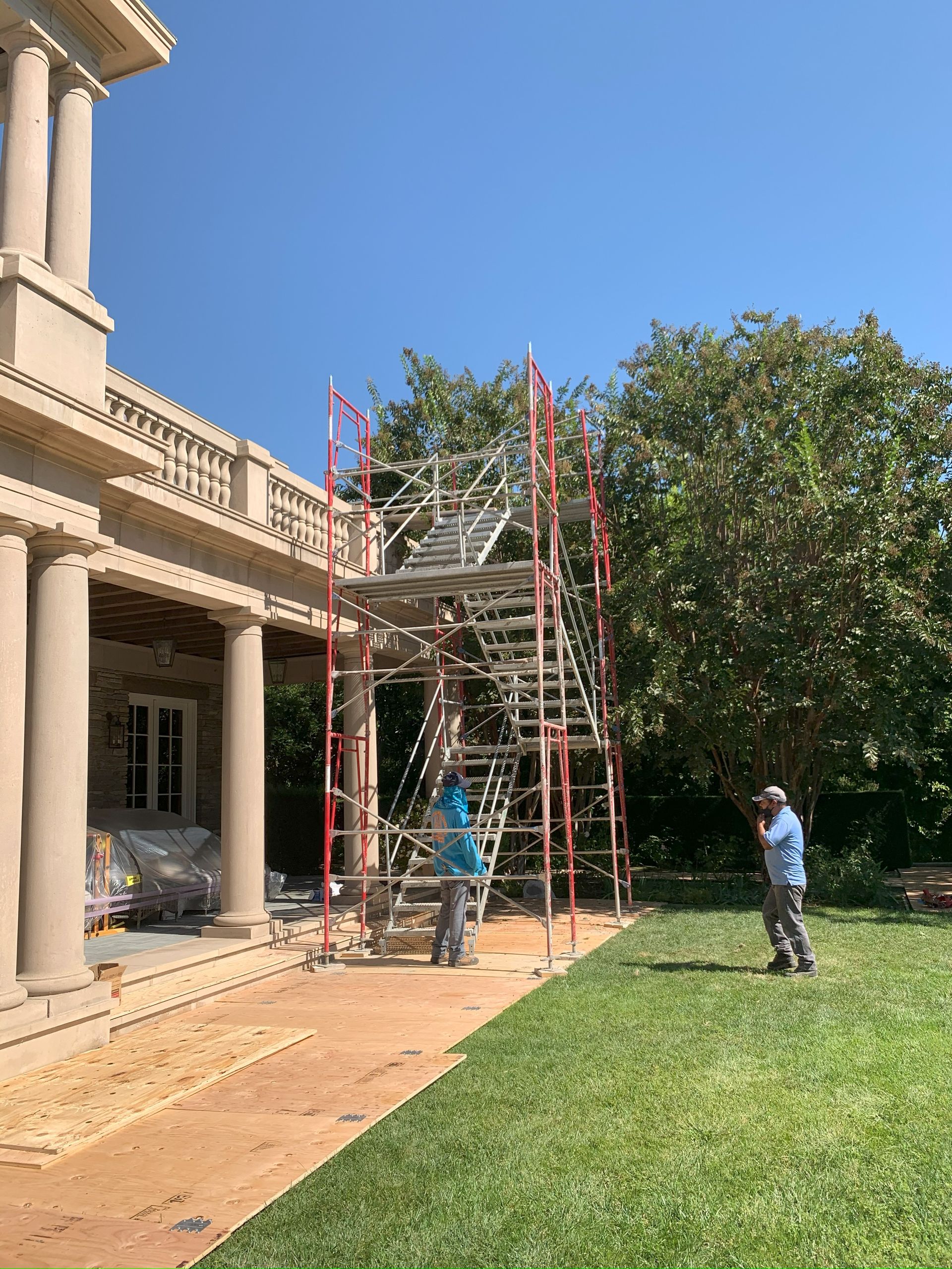 Scaffolding next to a beige house with columns; two people are working near the structure on a sunny day.