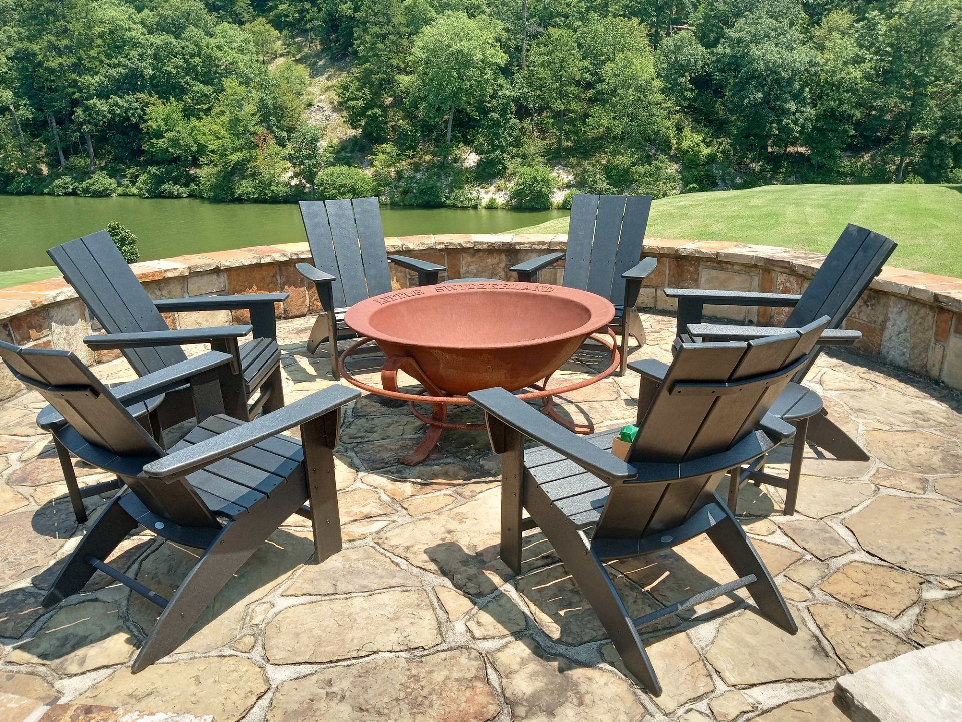 Six dark Adirondack chairs around a rusty fire pit on a stone patio overlooking a lake.