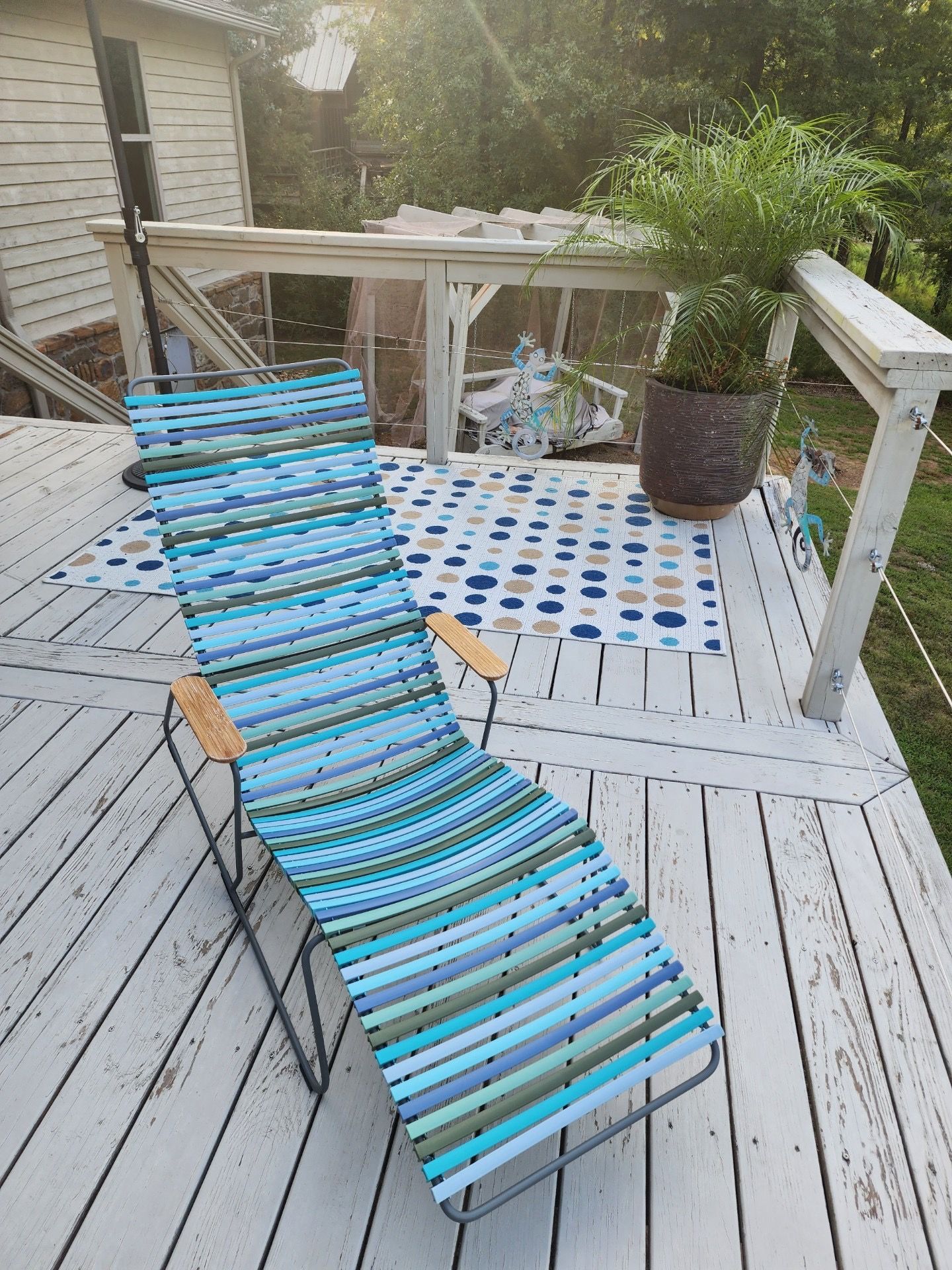 Lounge chair on a sunlit deck with a rug, potted plant, and weathered white railing.