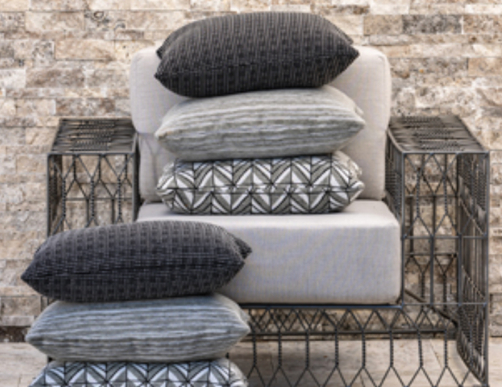 A chair with multiple grey and patterned throw pillows stacked on the seat and floor in front of a brick wall.