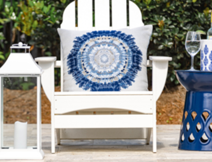 White Adirondack chair with blue mandala pillow, lantern, and side table. Outdoor setting.