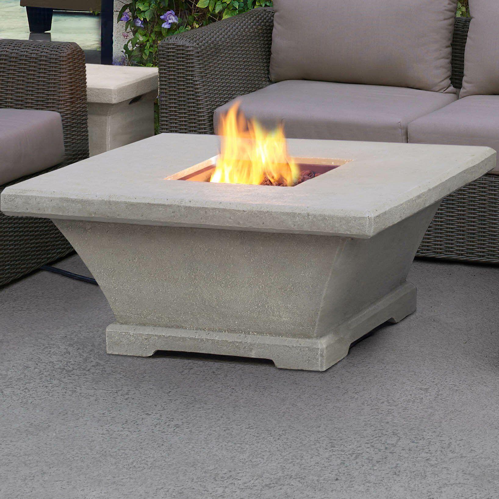 A lit square concrete fire pit on a patio, with a couch and other furniture in the background.