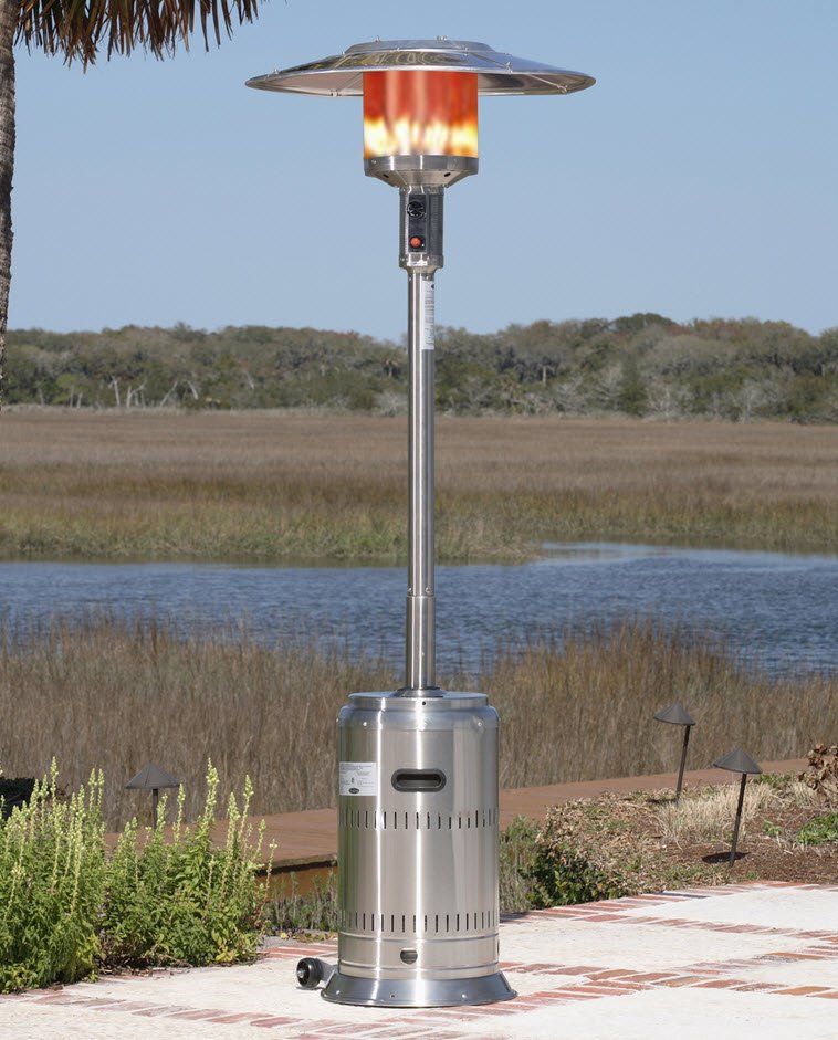 A tall, silver outdoor patio heater with flames lit, near a body of water.