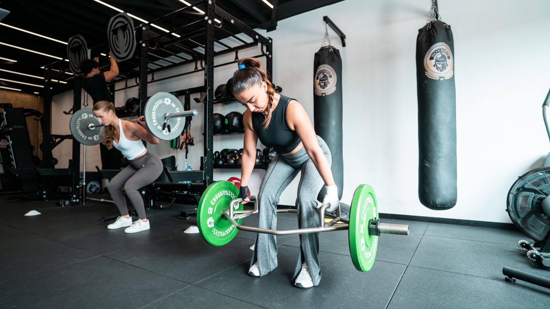 Two people lifting weights in a gym. One uses a barbell, the other a hex bar, both with green weights.