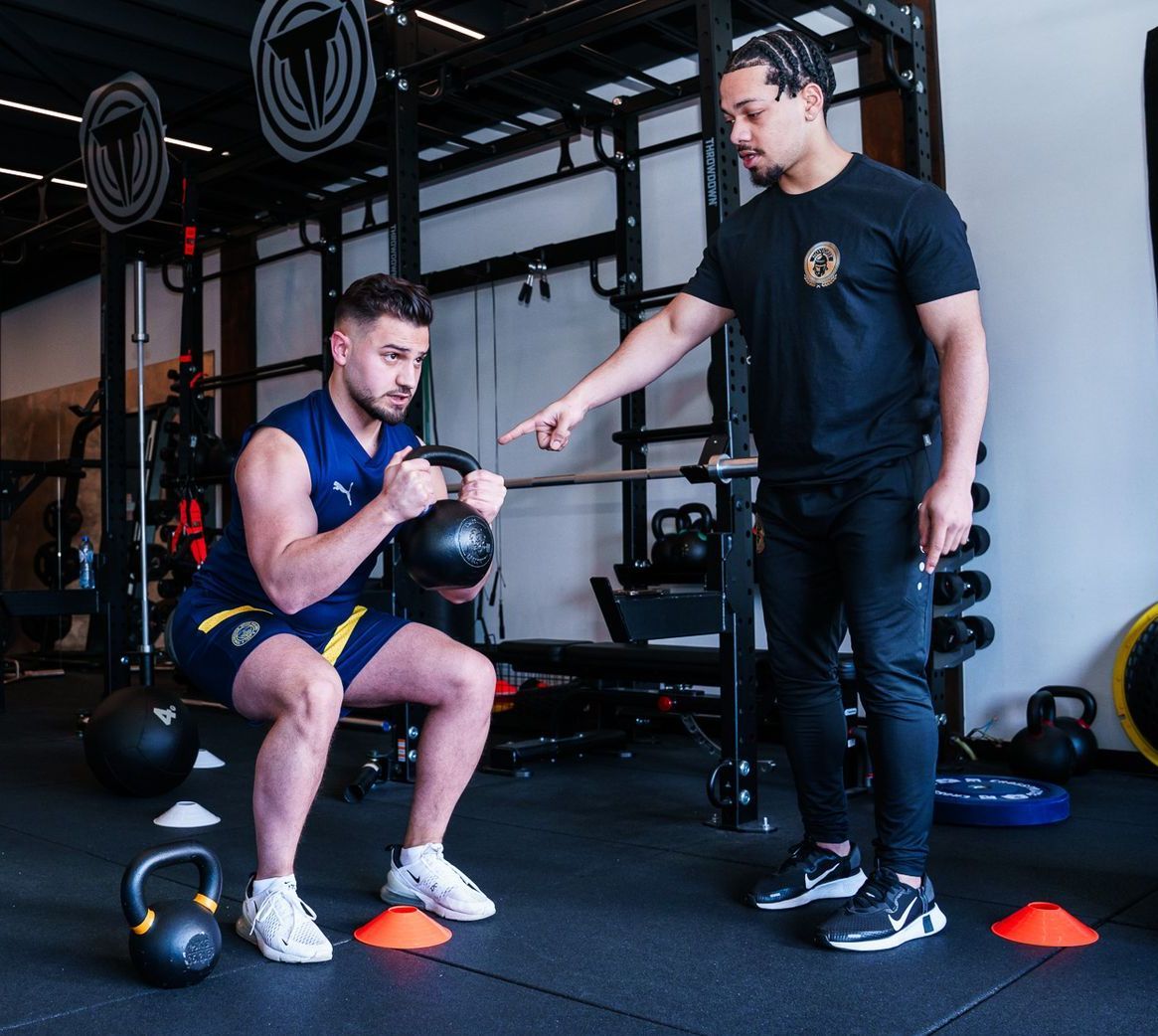 Een man hurkt in een sportschool terwijl een andere man naar hem wijst.