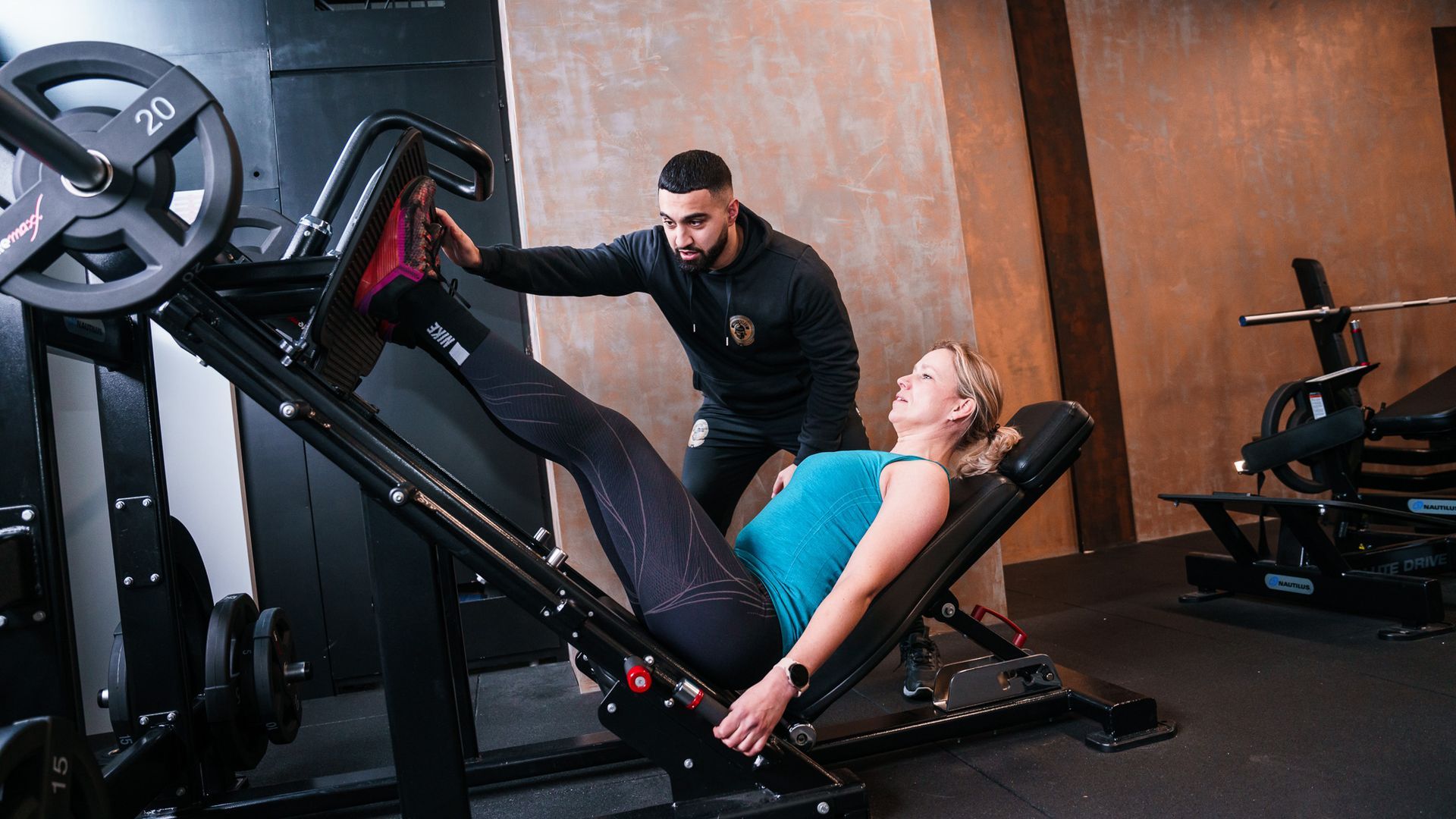 Een man helpt een vrouw met beenpressoefeningen in een sportschool.