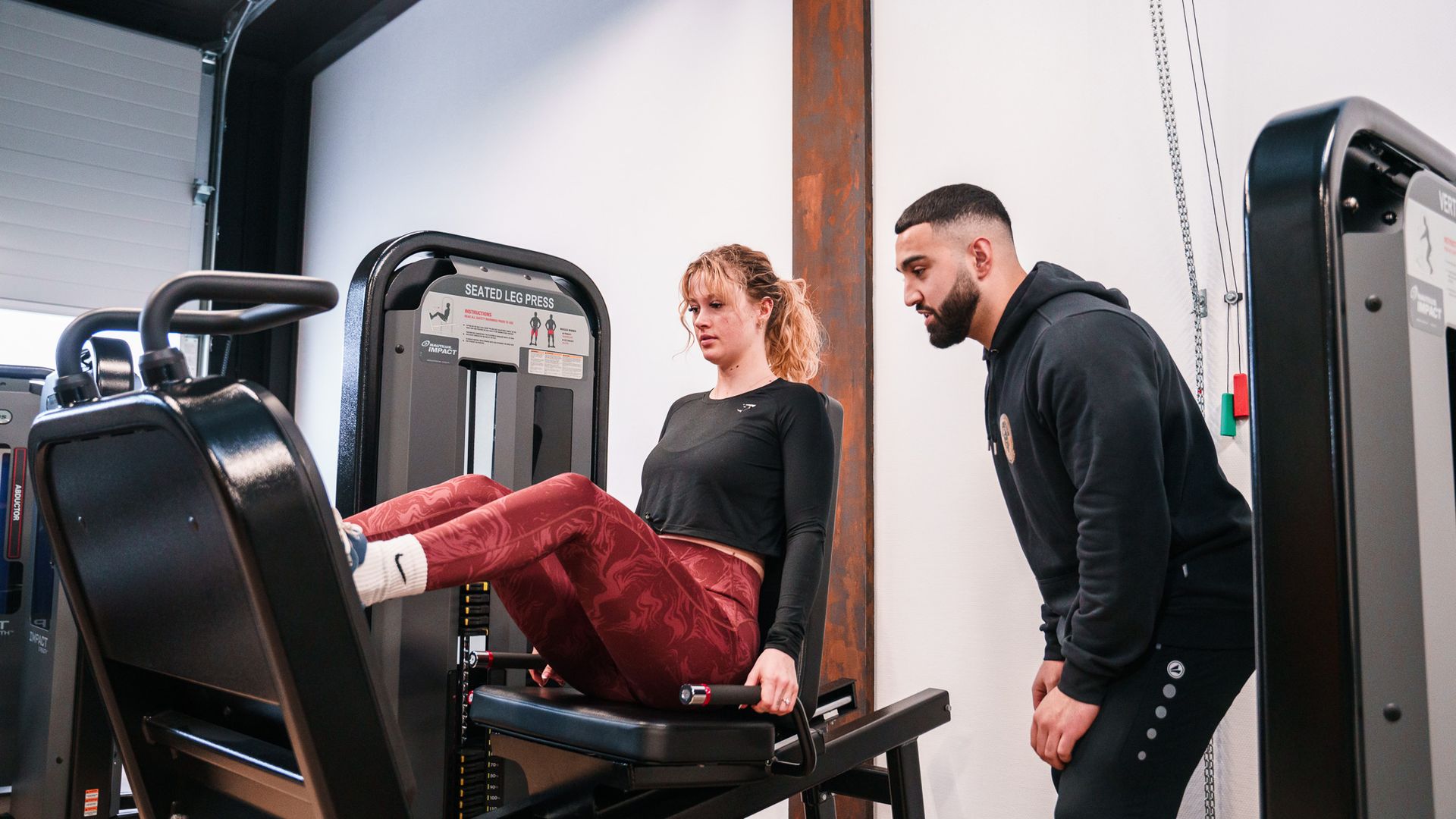Een man staat naast een vrouw die op een machine zit in een sportschool.