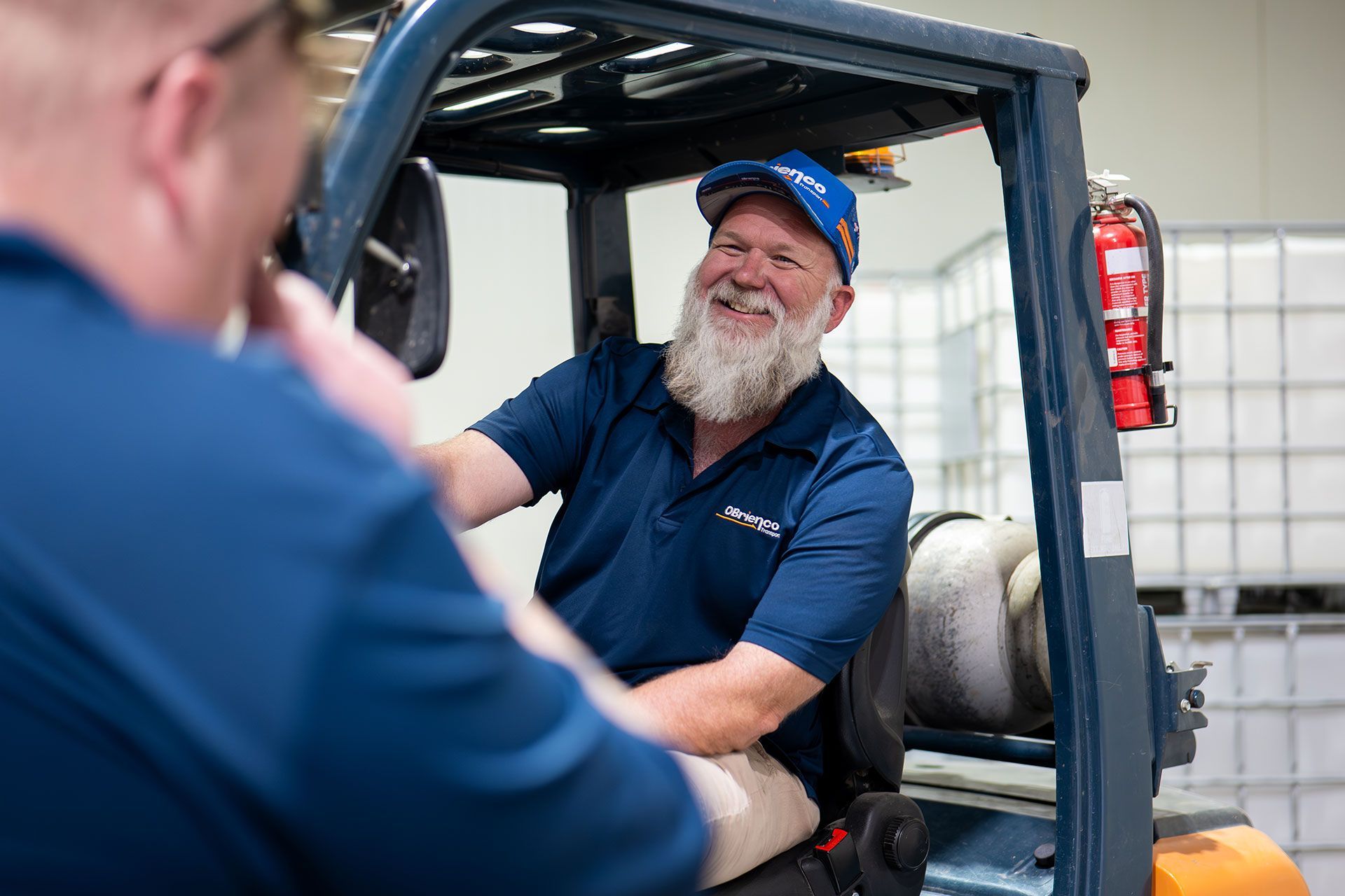 Man smiling, operating a forklift, talking to another person in a warehouse setting.
