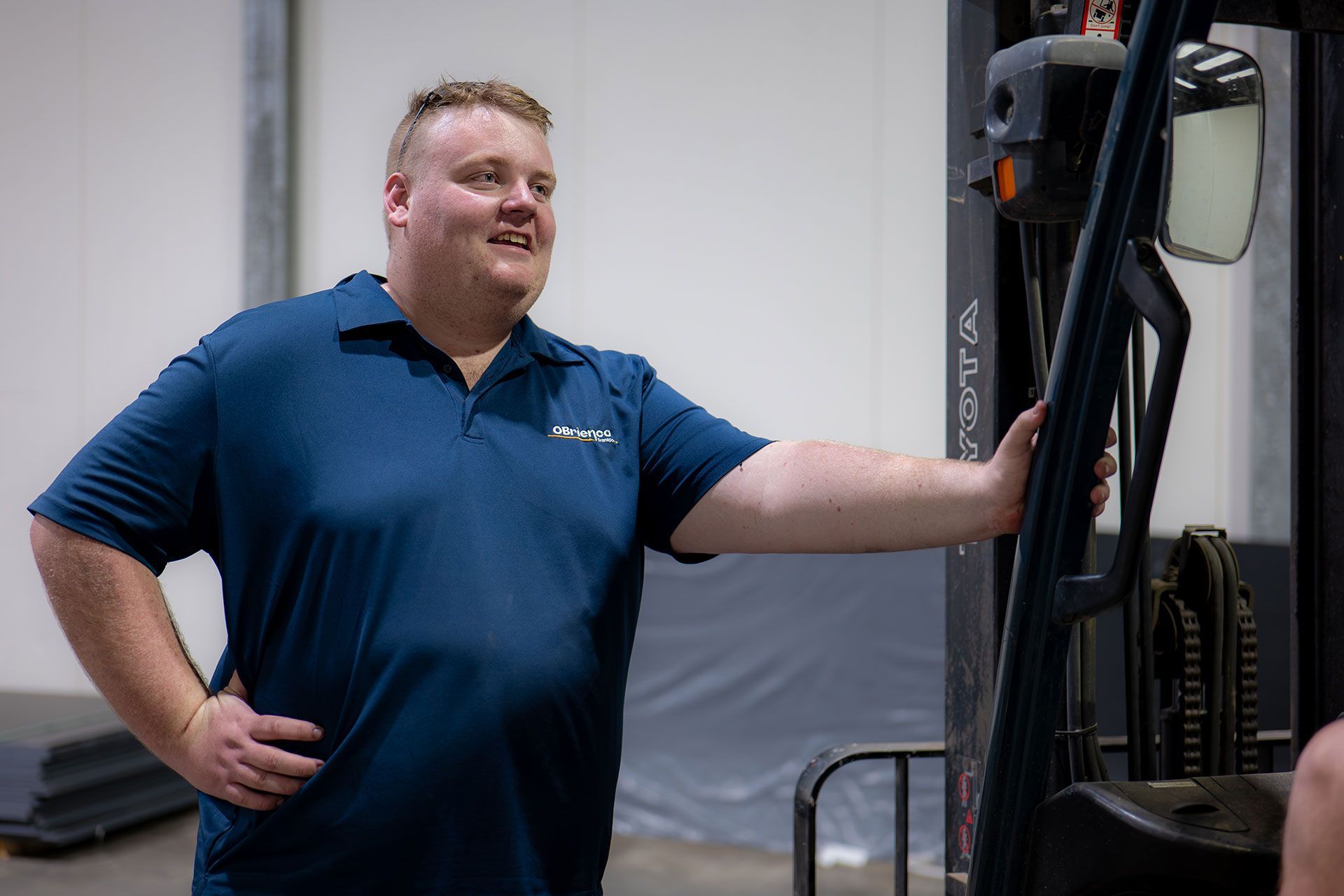 Man in blue shirt, smiling, leaning on a forklift in a warehouse.