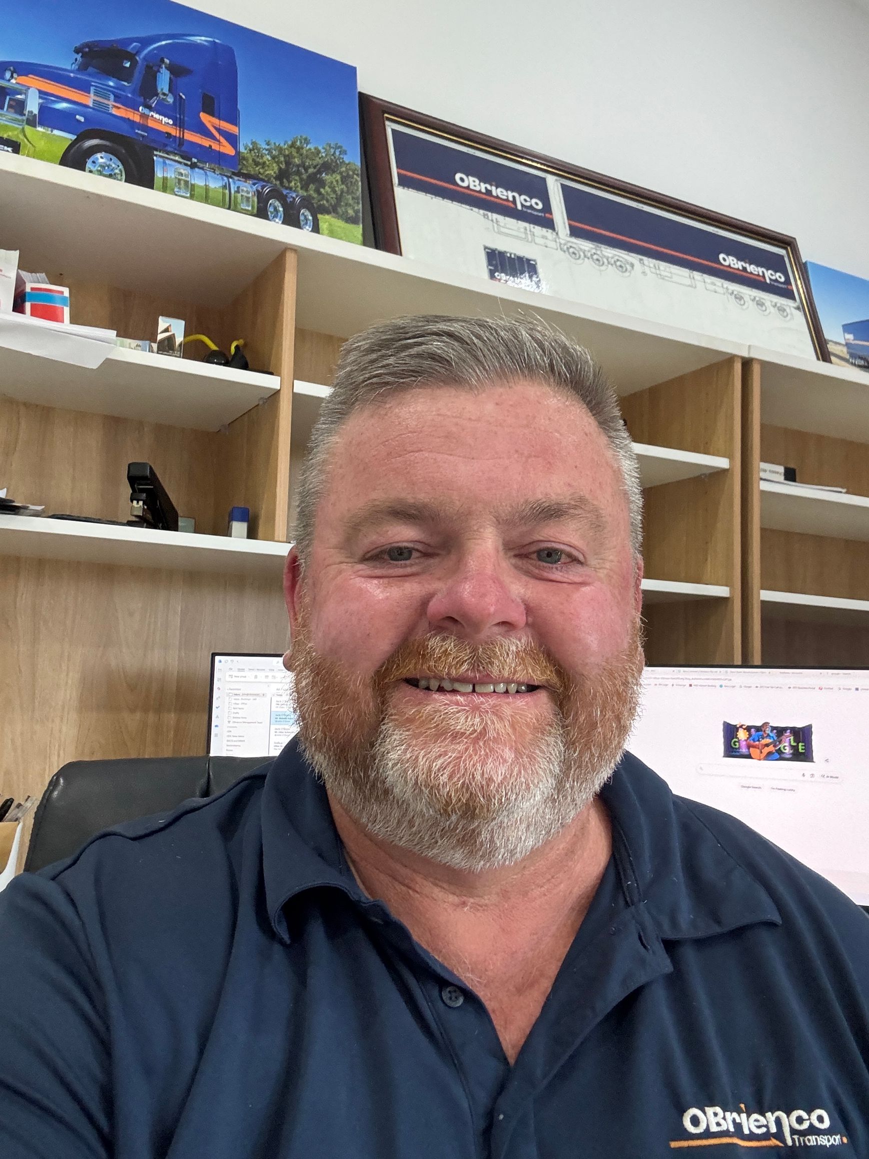 Man in a navy shirt smiles, sitting in an office with trucking-related artwork and shelves.
