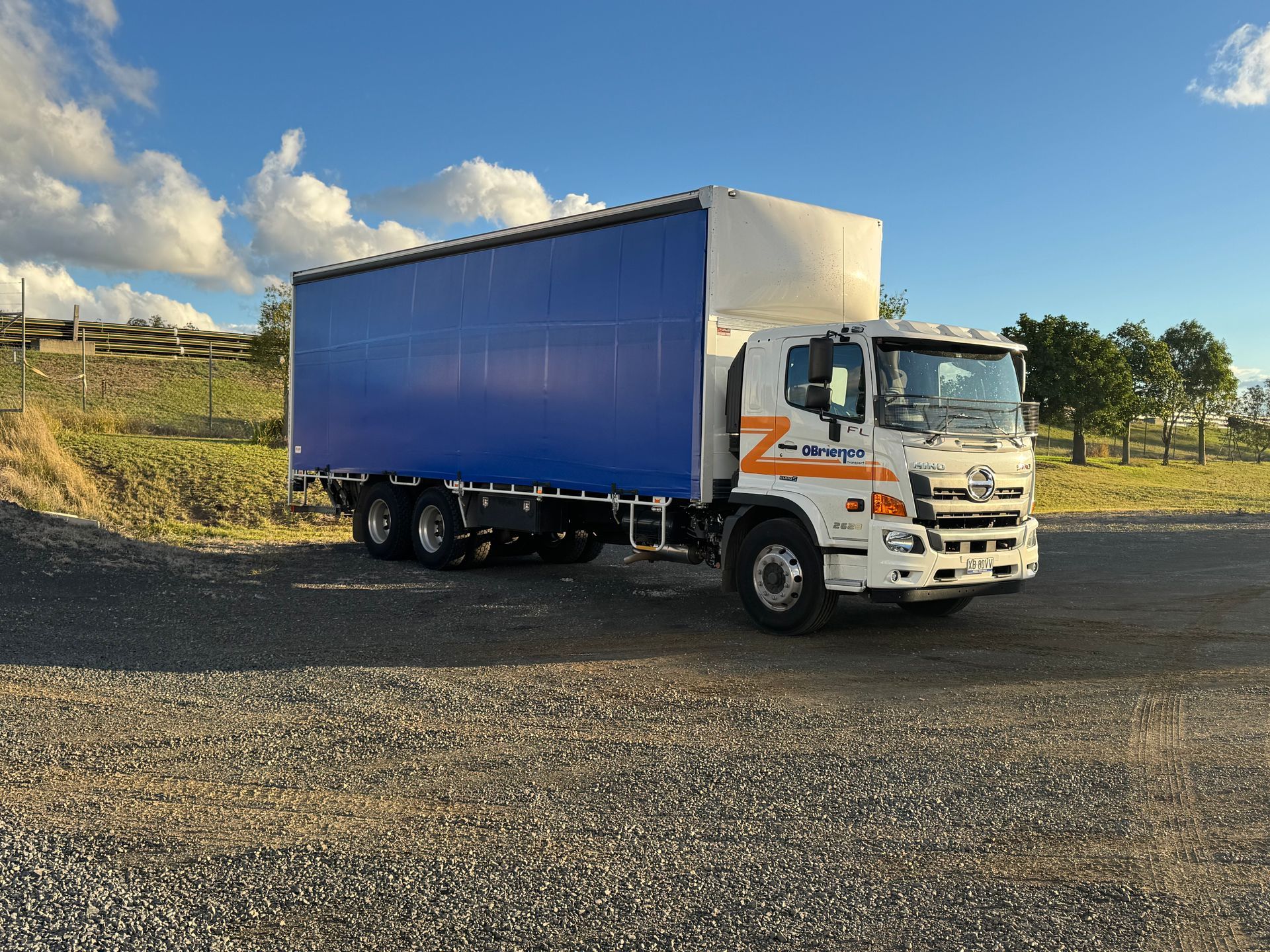 White and blue delivery truck parked on gravel in front of a grassy field under a cloudy sky.
