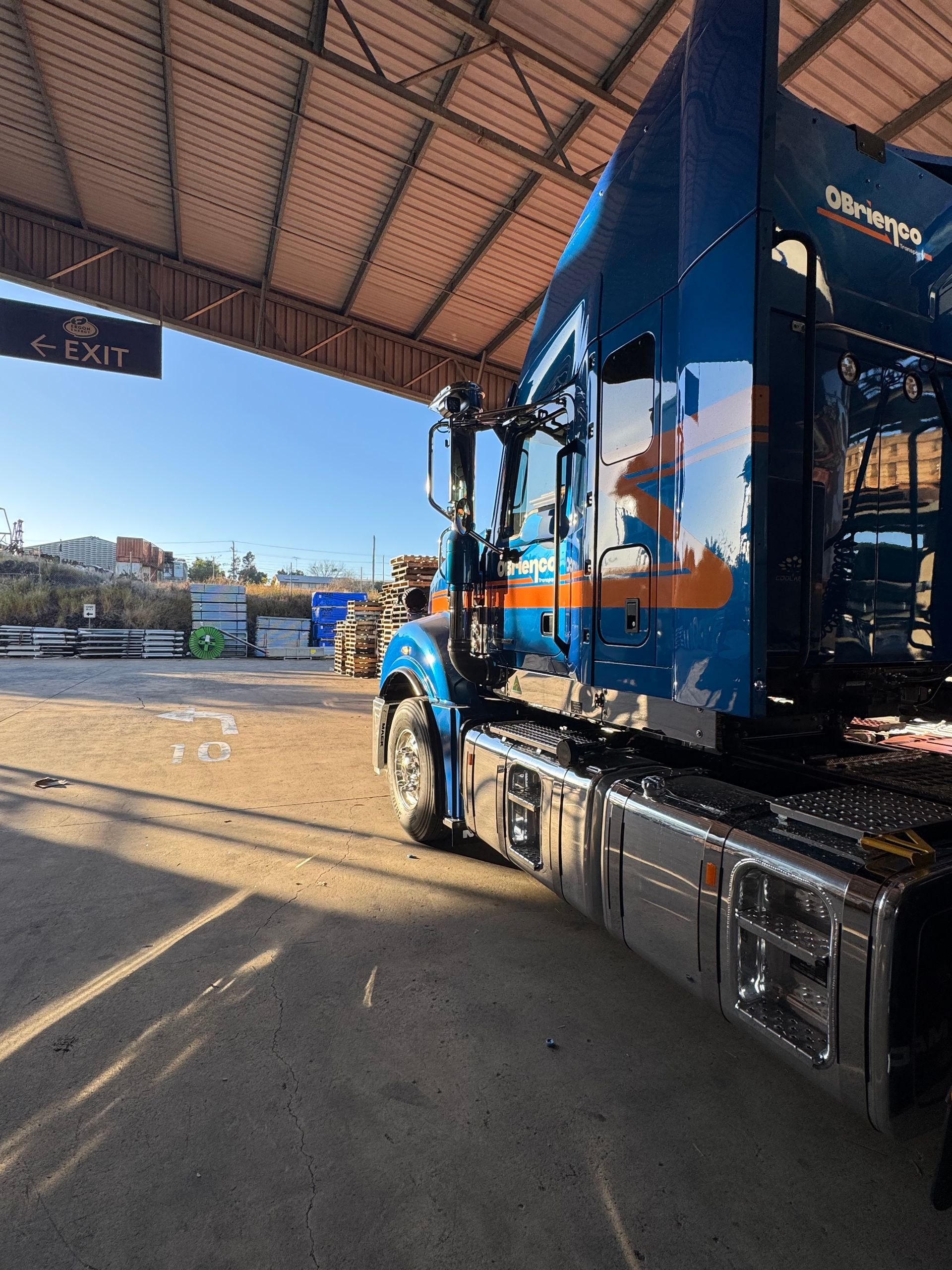 Blue semi-truck parked under a shelter. The truck has orange and silver accents, with an open yard in the background.