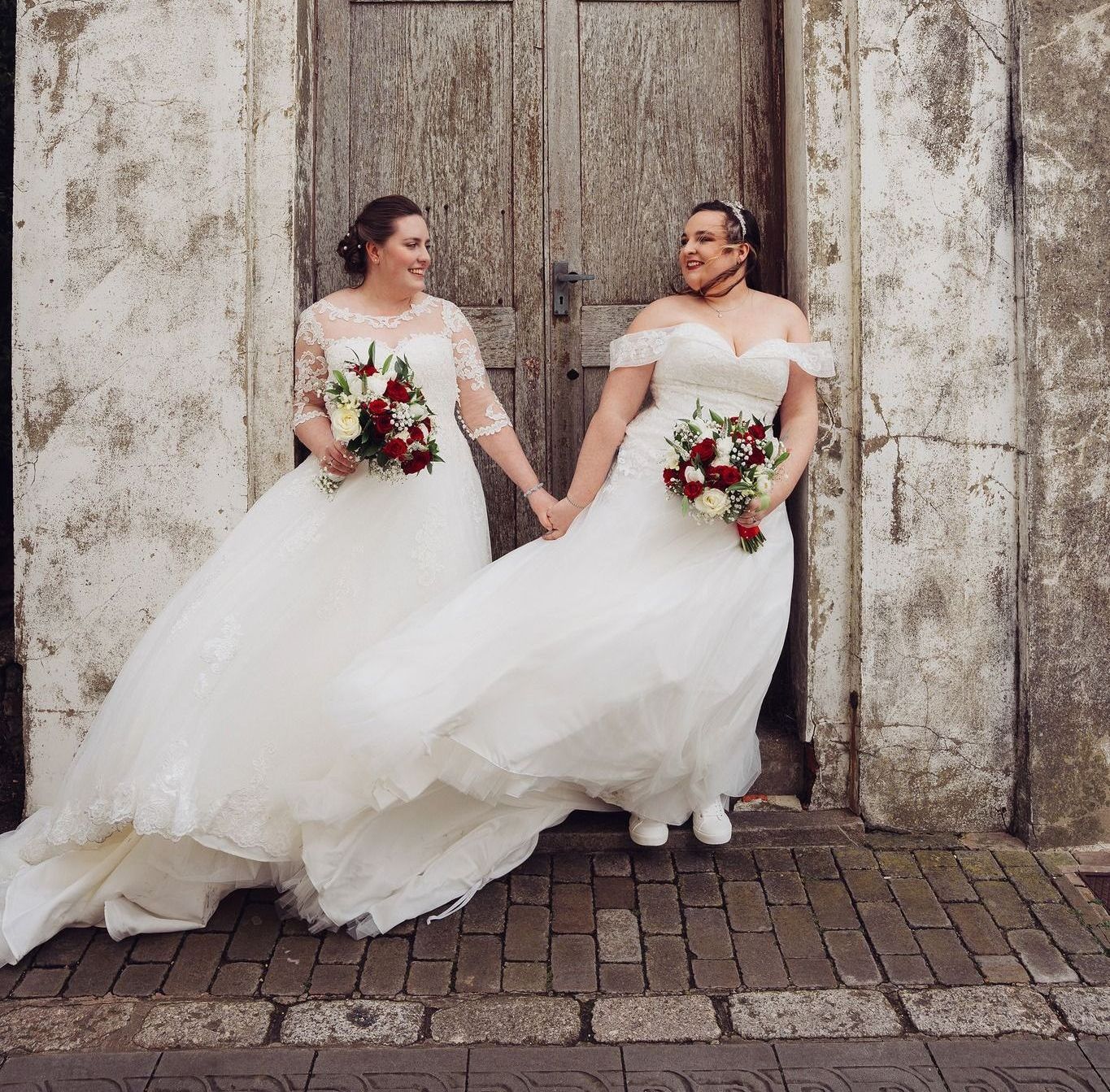 Two women in wedding dresses are standing next to each other holding hands.
