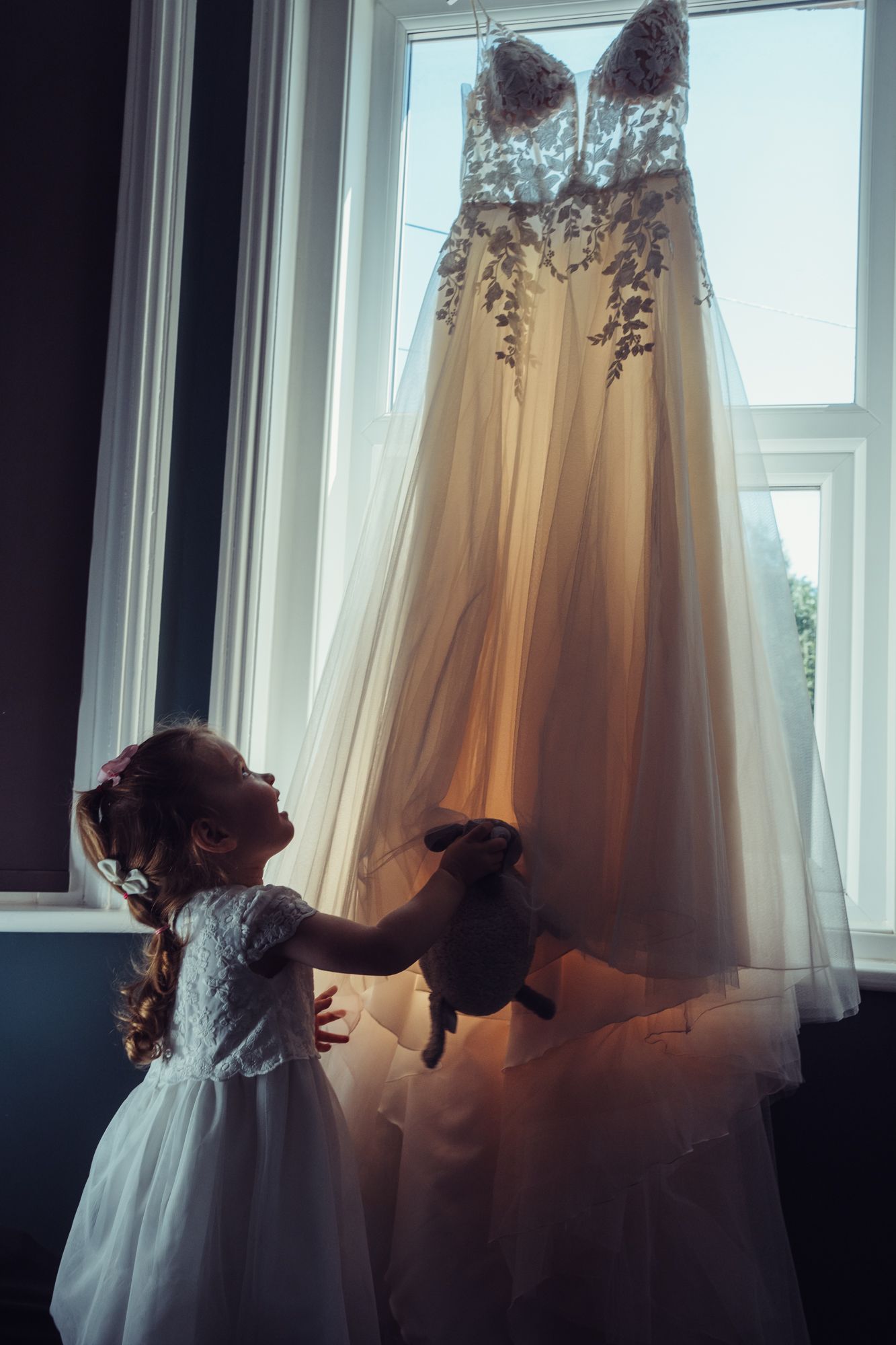 A little girl is holding a stuffed animal in front of a wedding dress hanging in a window.