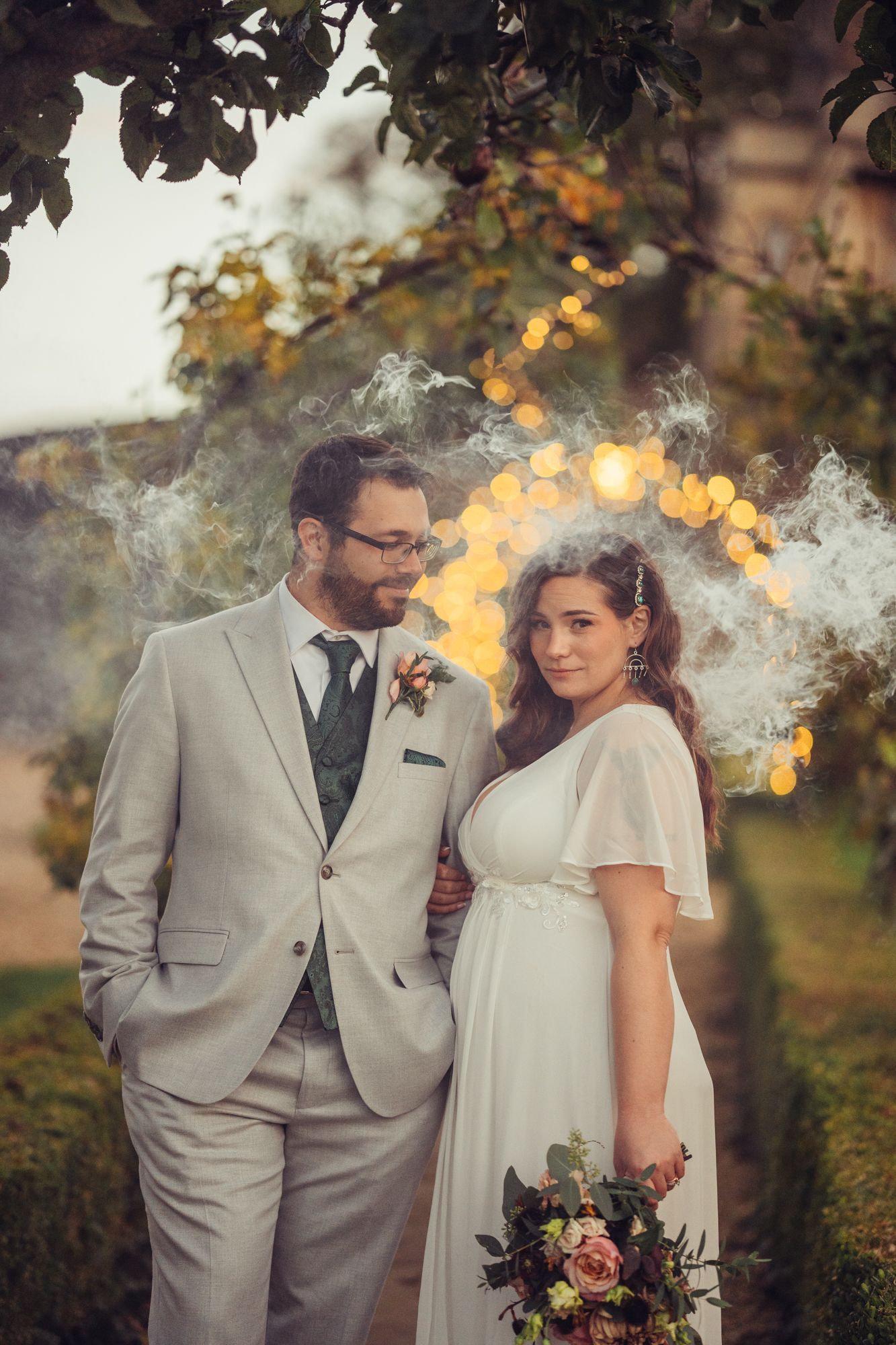 A bride and groom are posing for a picture in a garden with smoke coming out of their hair.