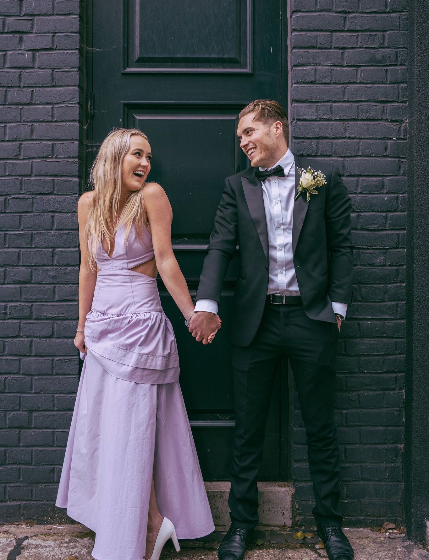 A man in a tuxedo and a woman in a purple dress are holding hands in front of a black door.