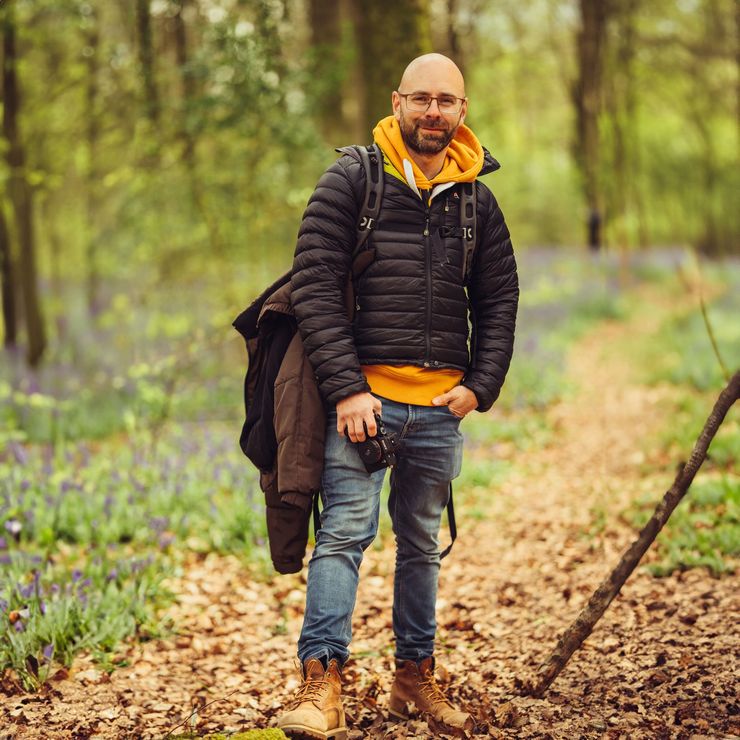 A man in a black jacket and orange hoodie is standing in the woods.