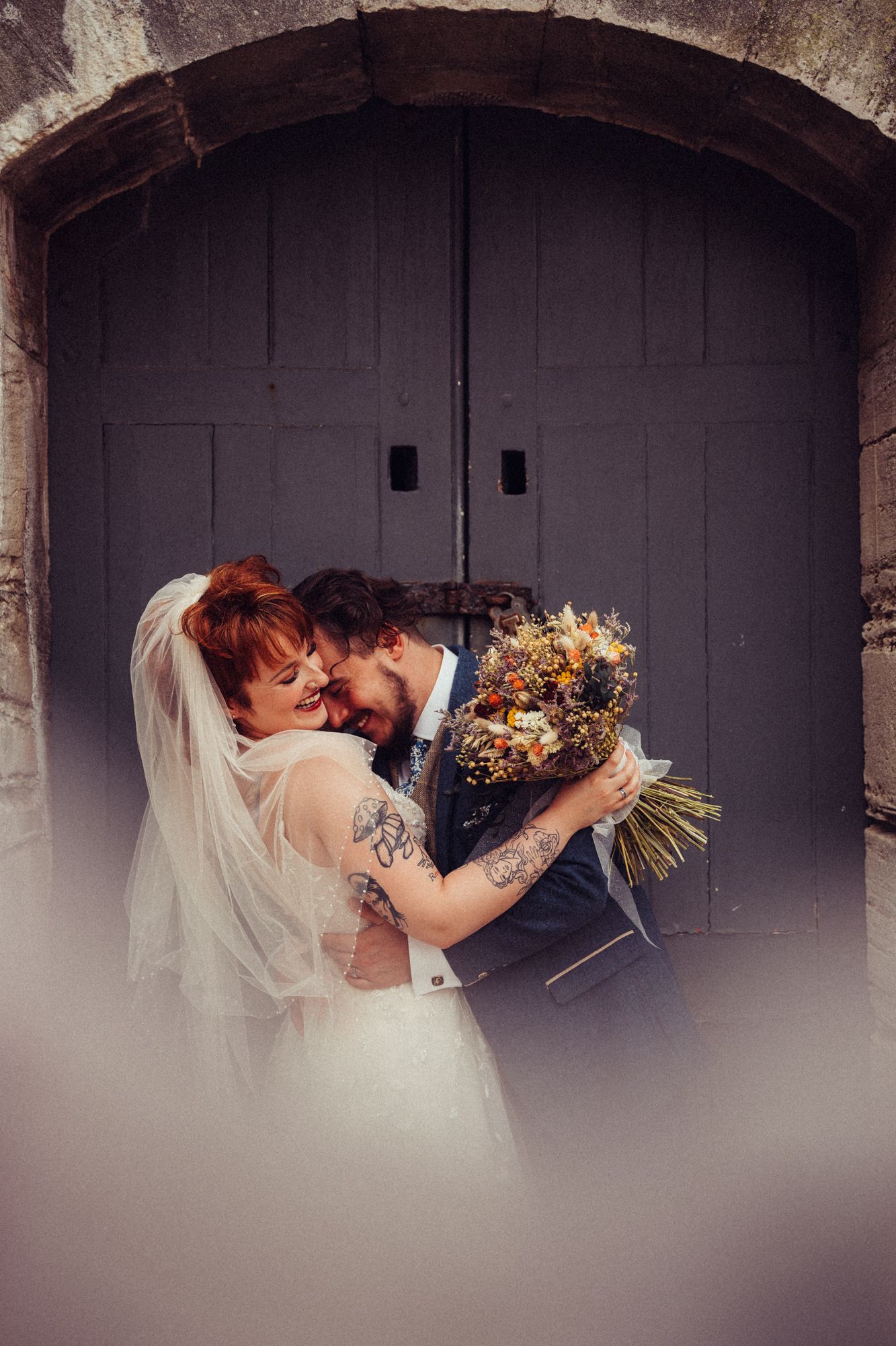 A bride and groom are hugging in front of a door . the bride is holding a bouquet of flowers.