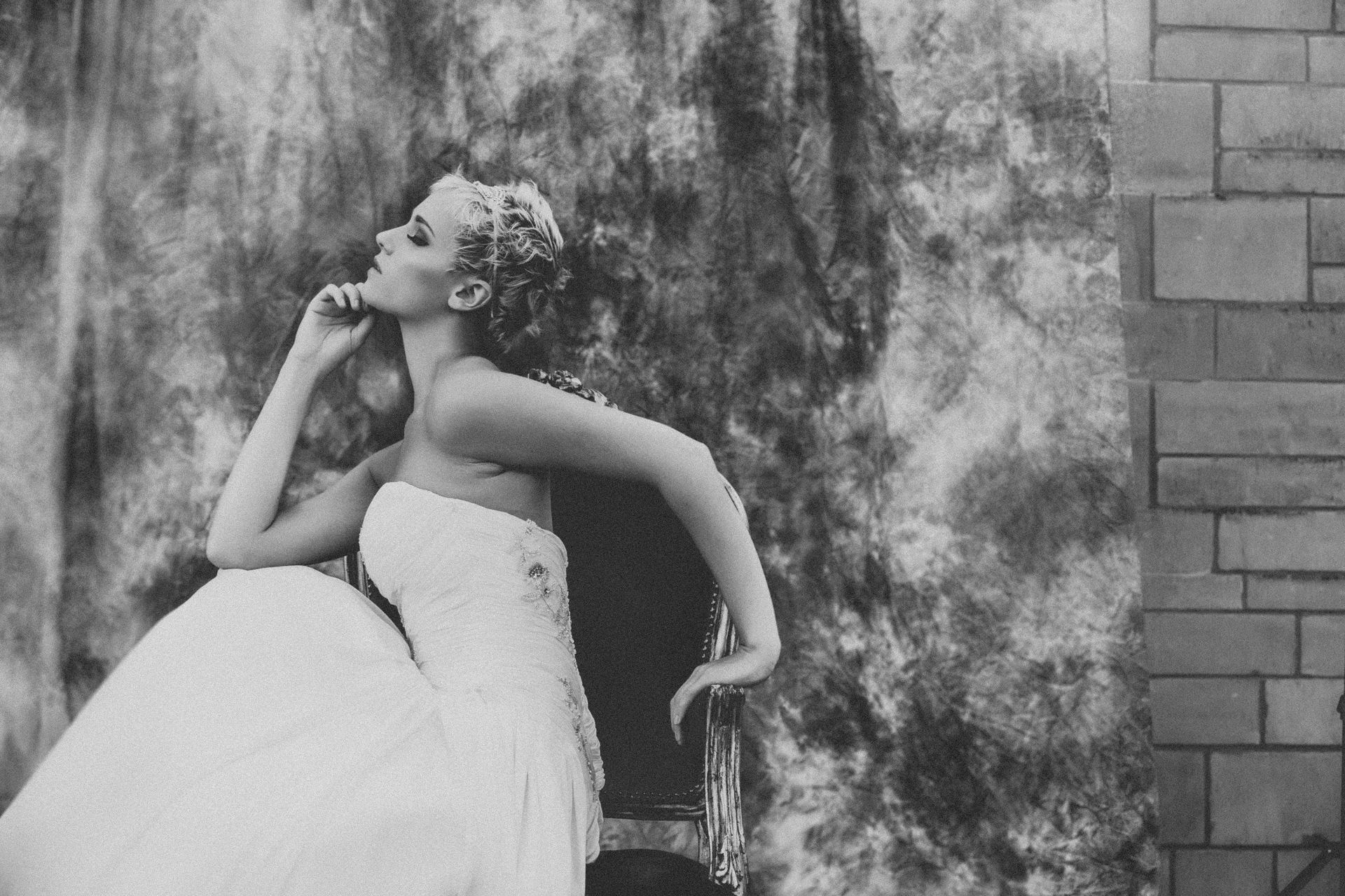 A woman in a wedding dress is sitting in a chair in front of a brick wall.