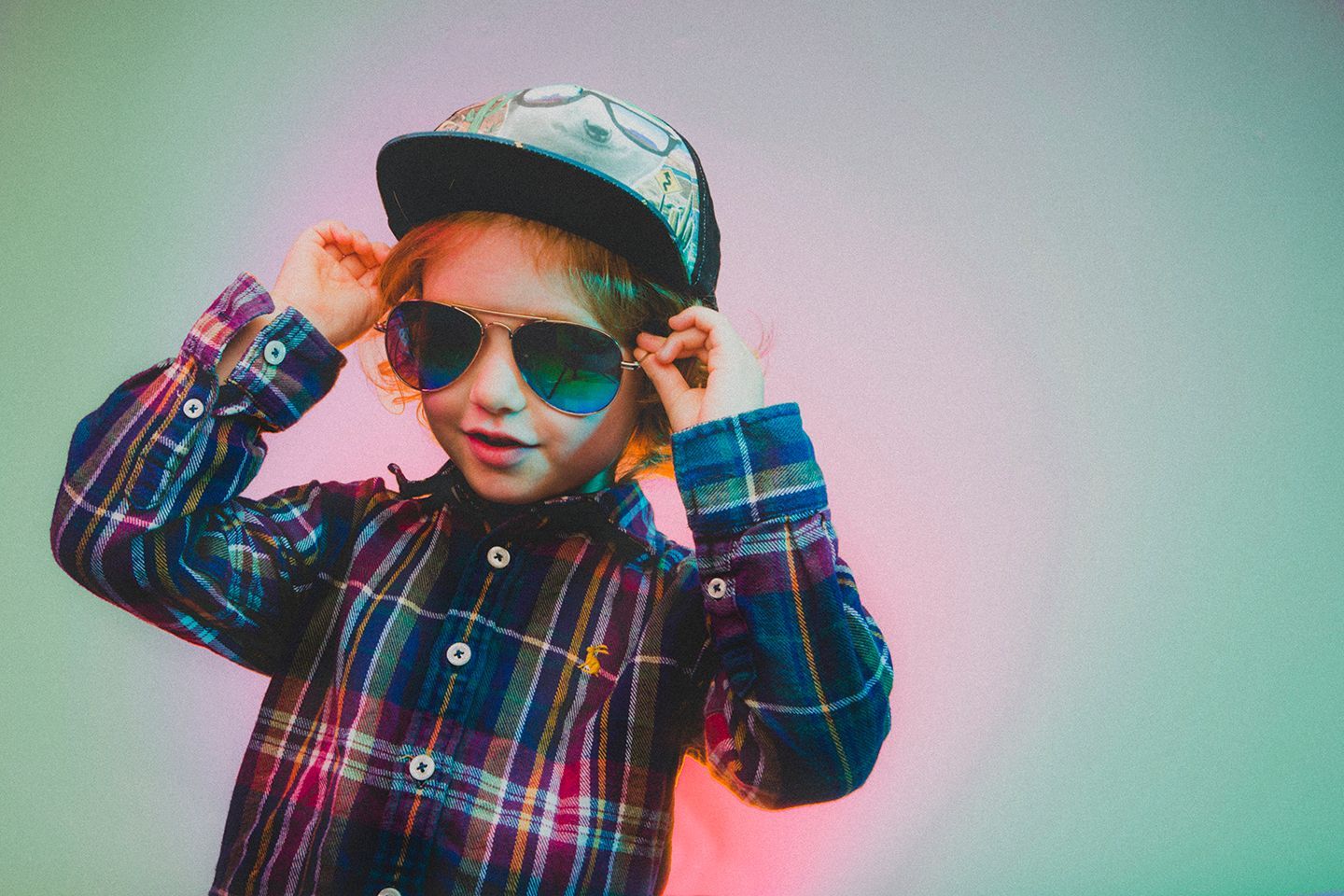A young boy wearing sunglasses and a hat is adjusting his sunglasses.