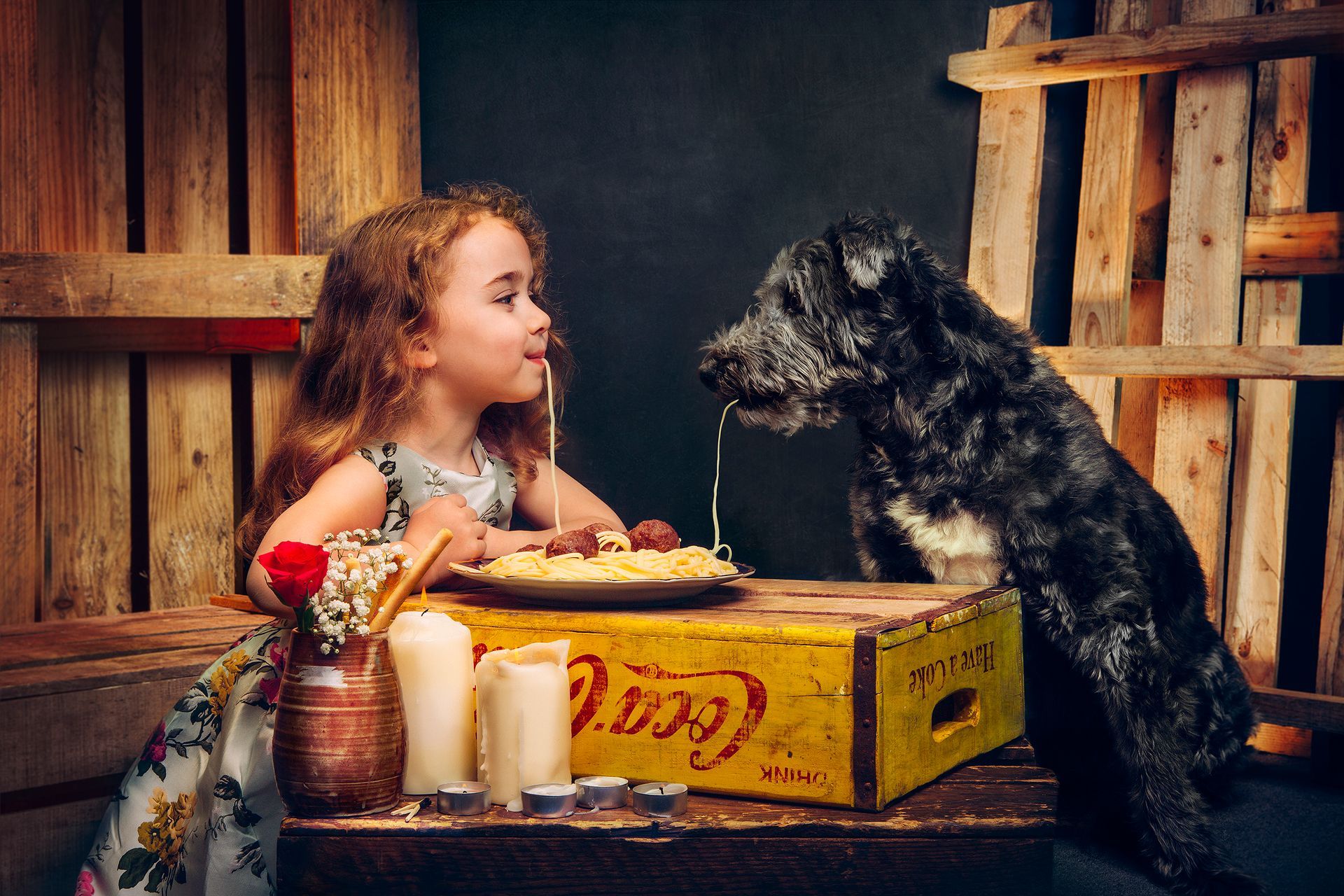 A little girl is sitting at a table with a dog and a plate of food.