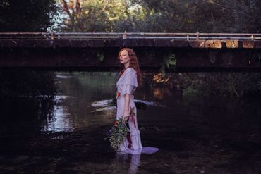 A woman in a white dress is standing in a river under a bridge.