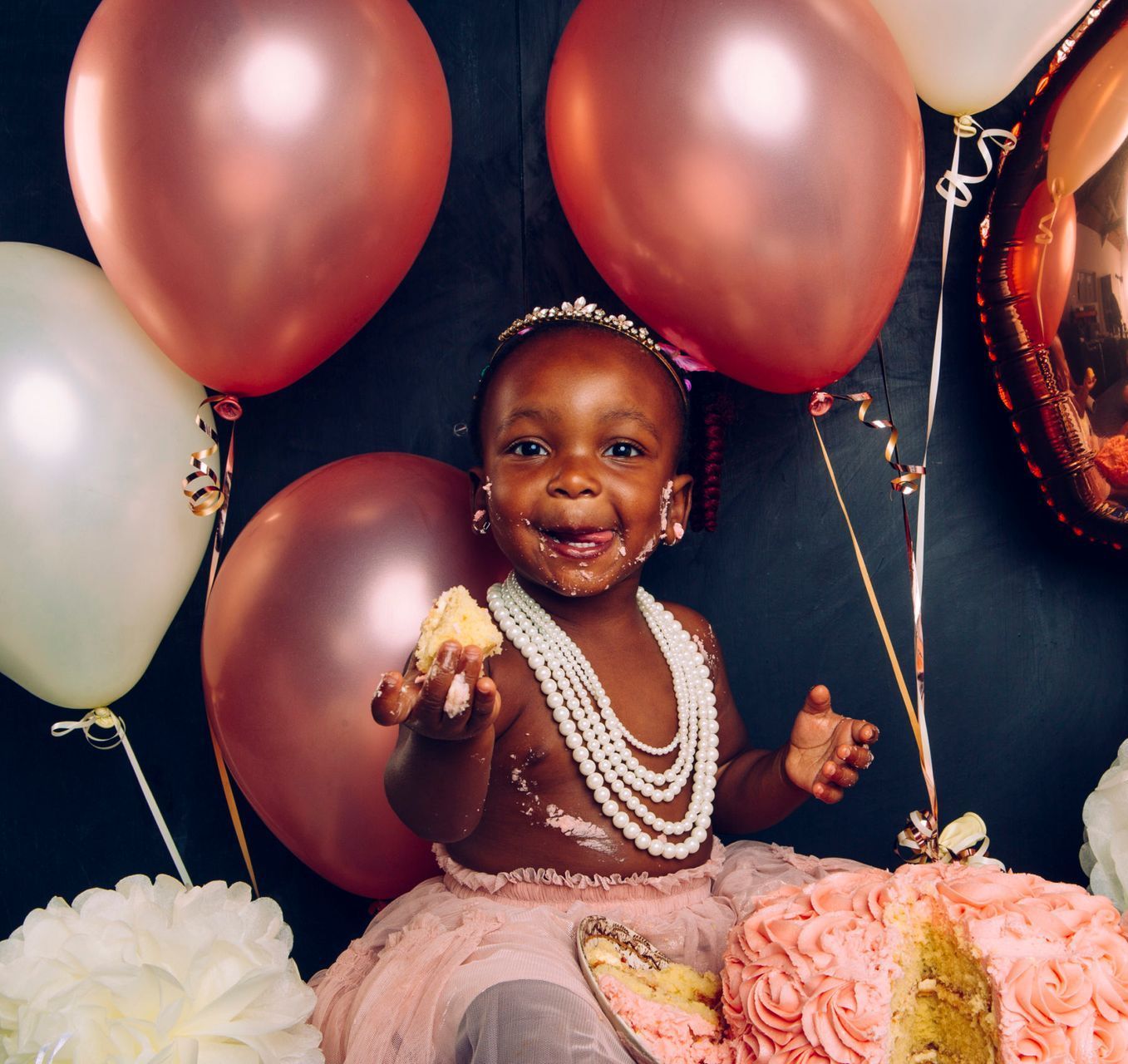 A little girl is sitting next to a cake and balloons