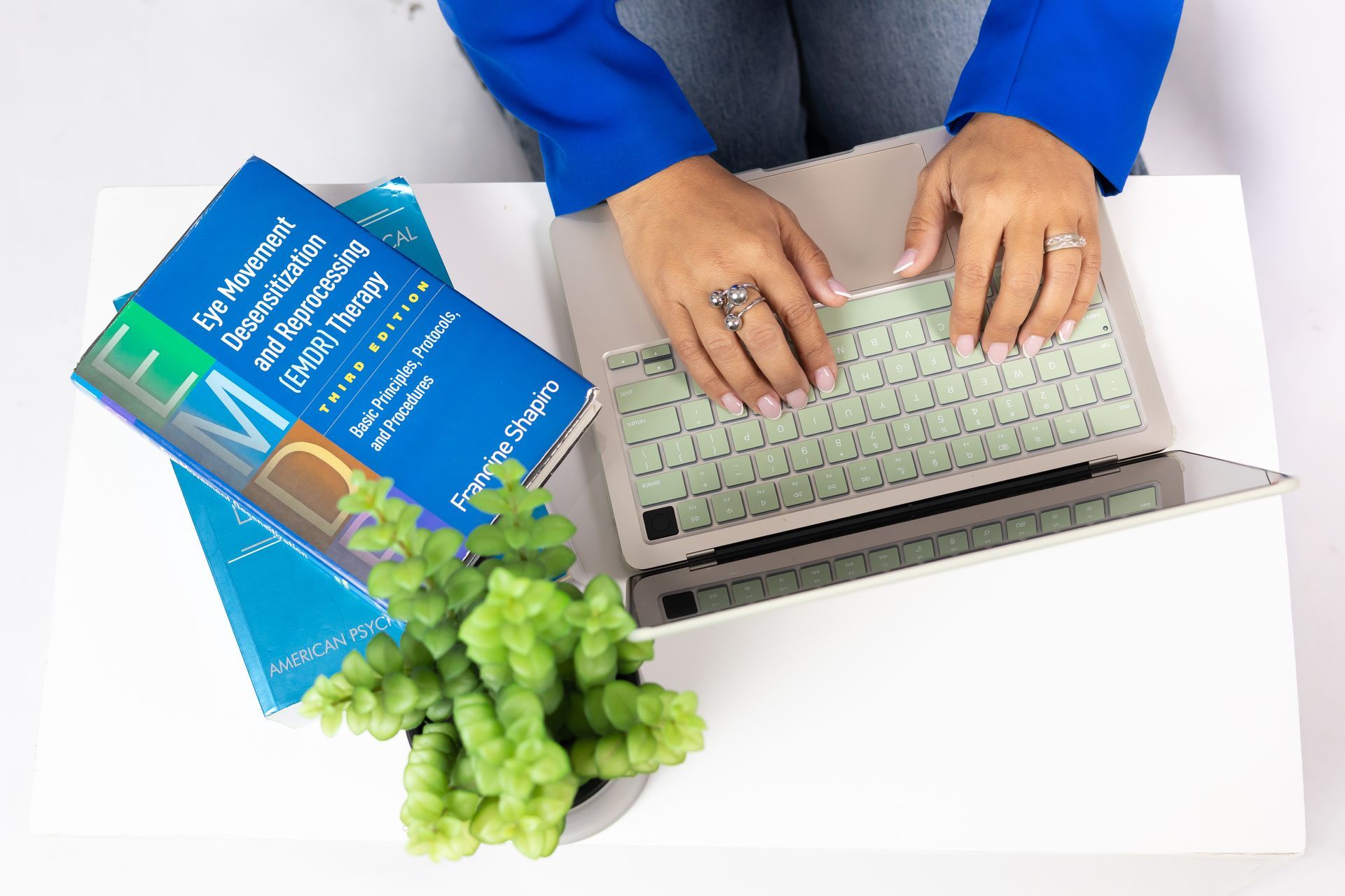 Hands typing on a laptop at a white desk with books and a small plant.