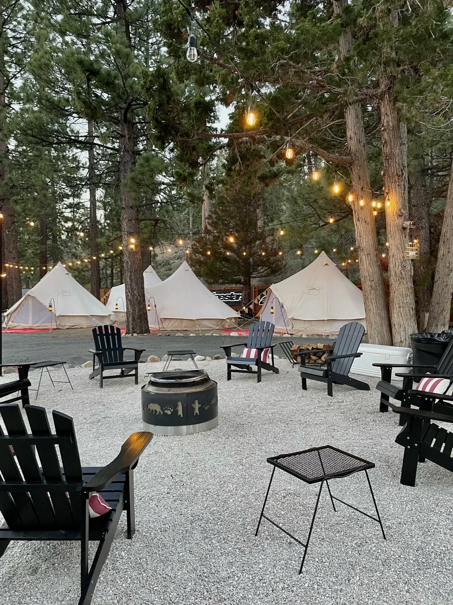 Campground with tents, string lights, a fire pit, and Adirondack chairs surrounded by trees.
