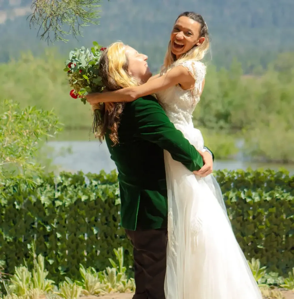 Man in green coat lifts bride in white dress, both smiling, outdoors.