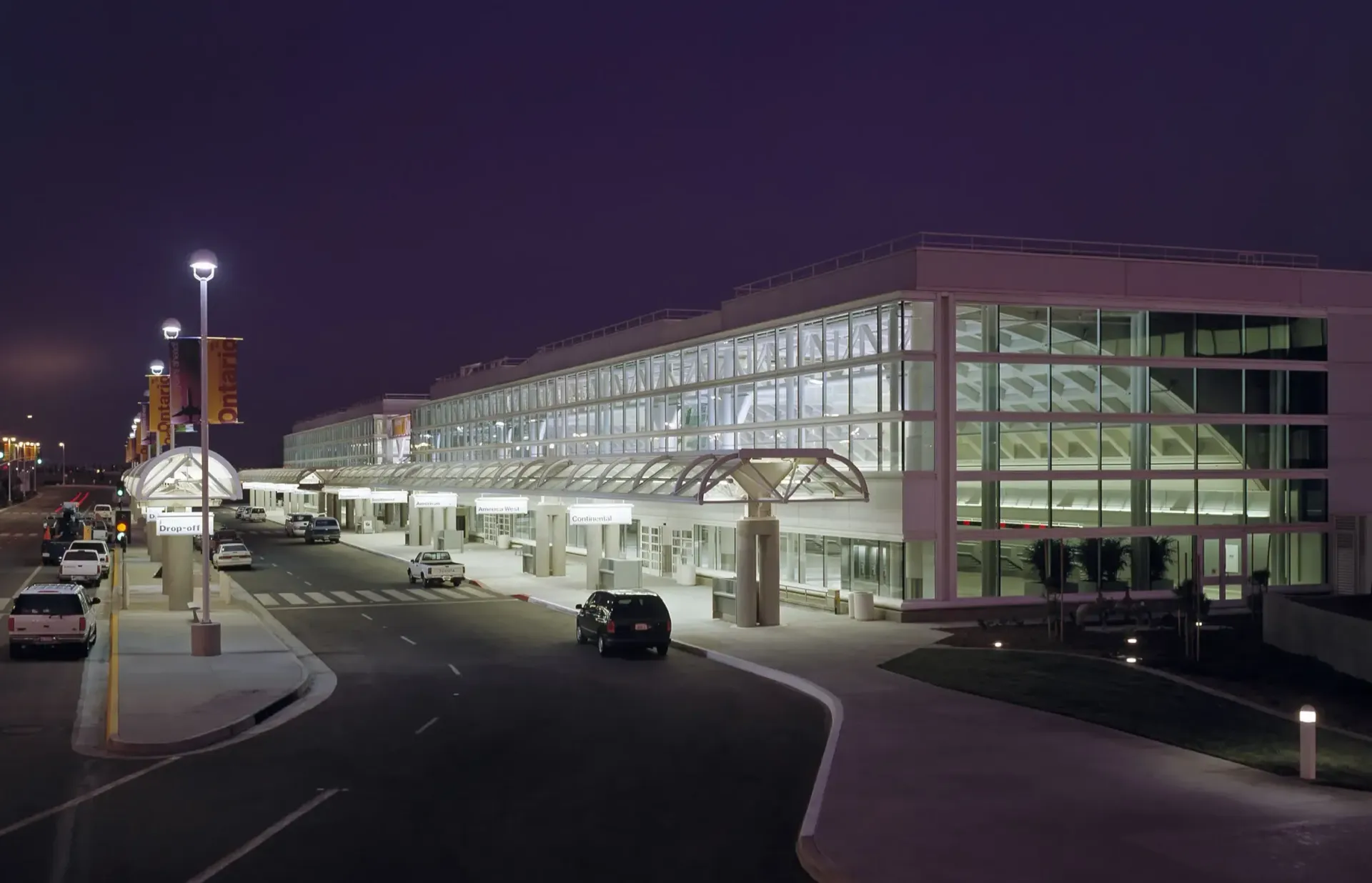 Airport terminal exterior at dusk, illuminated with lights, cars on roadway, overcast sky.