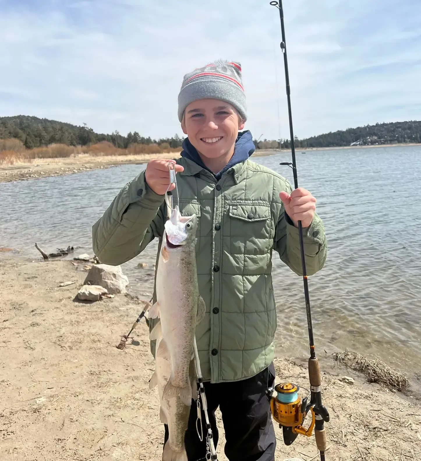 Boy holding a caught fish by a lake, wearing a green jacket and beanie, smiling.