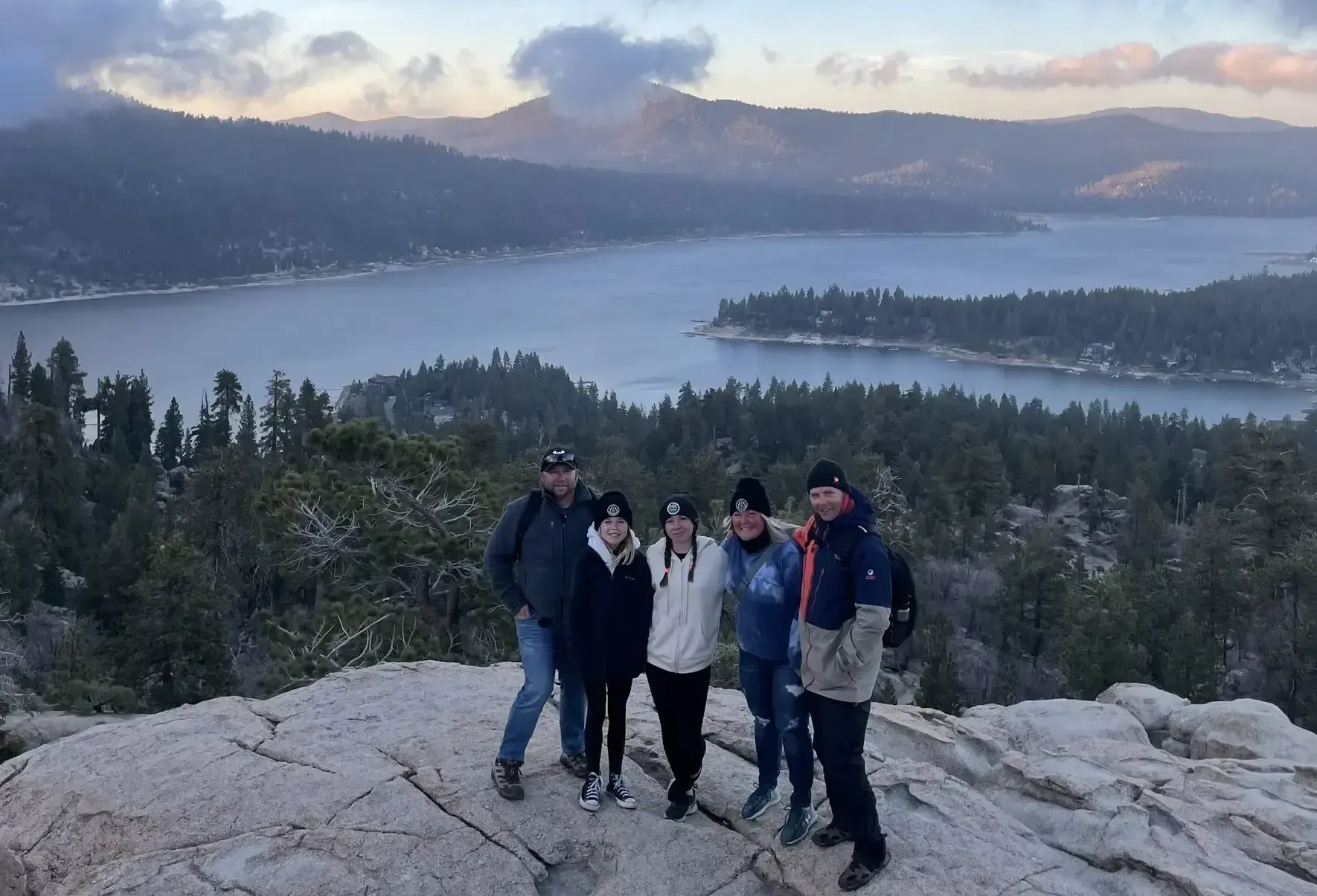Group of five people posing on a rocky mountaintop overlooking a lake and mountains.