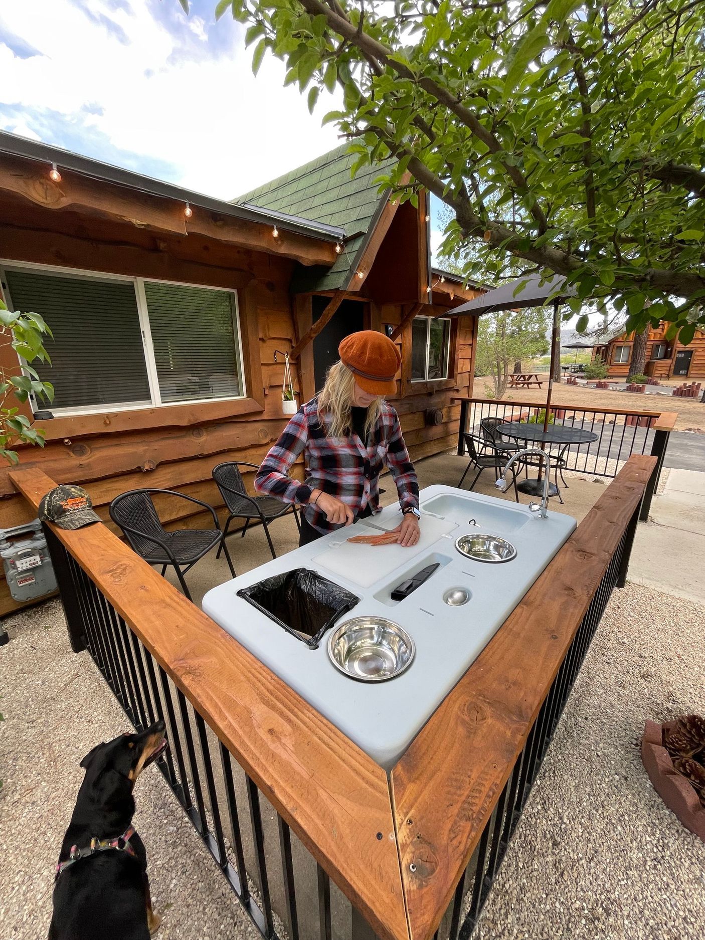Woman in orange hat prepares food at outdoor counter, dog watches. Wooden deck, cabin background.