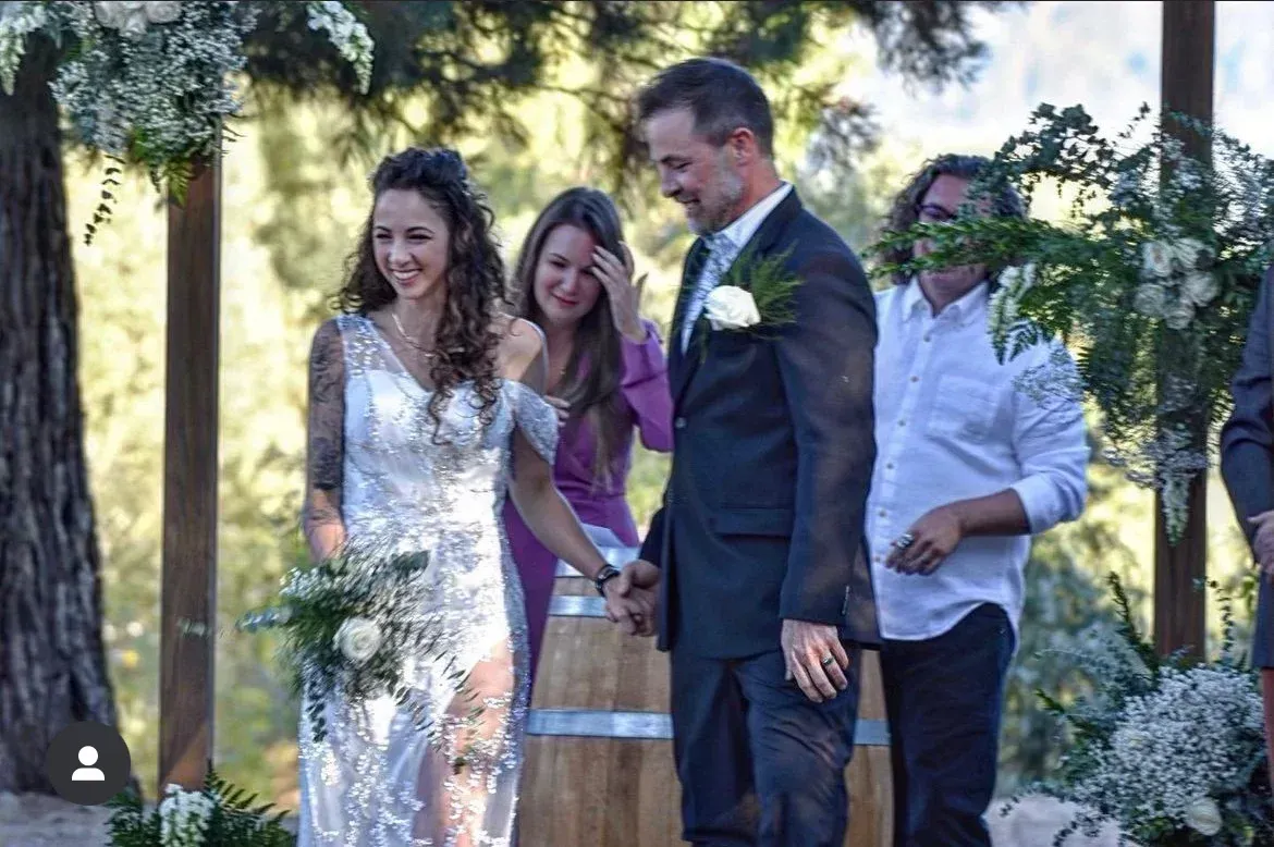 Bride and groom holding hands during outdoor wedding ceremony; floral arch, smiling, happy expressions.